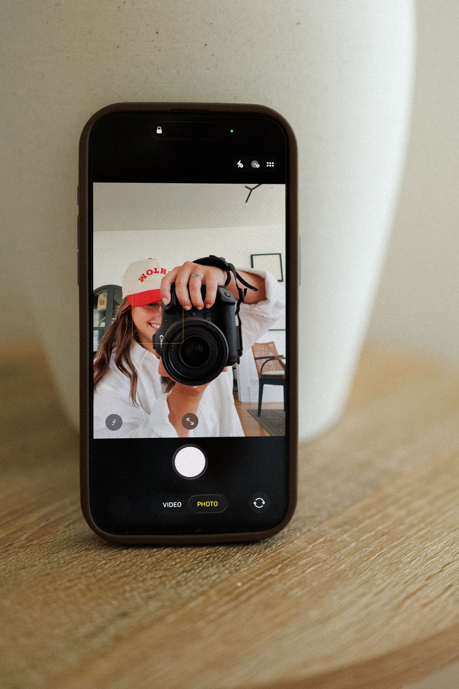 Phone screen showing a mirror selfie of a person in a red cap holding a DSLR camera.
