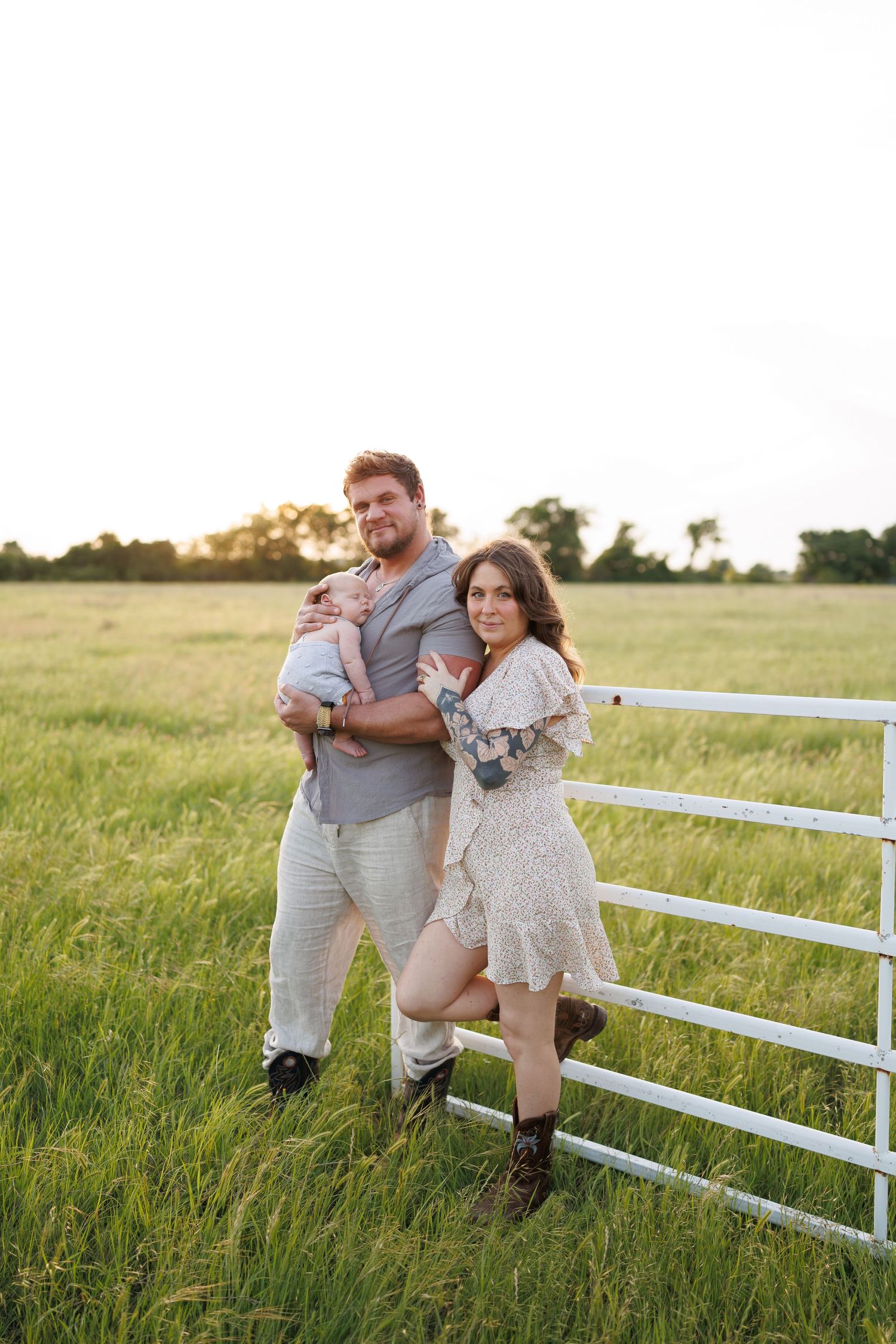 A young family poses by a white farm gate in a lush green field at golden hour, man holding a baby beside a woman in a floral dress.