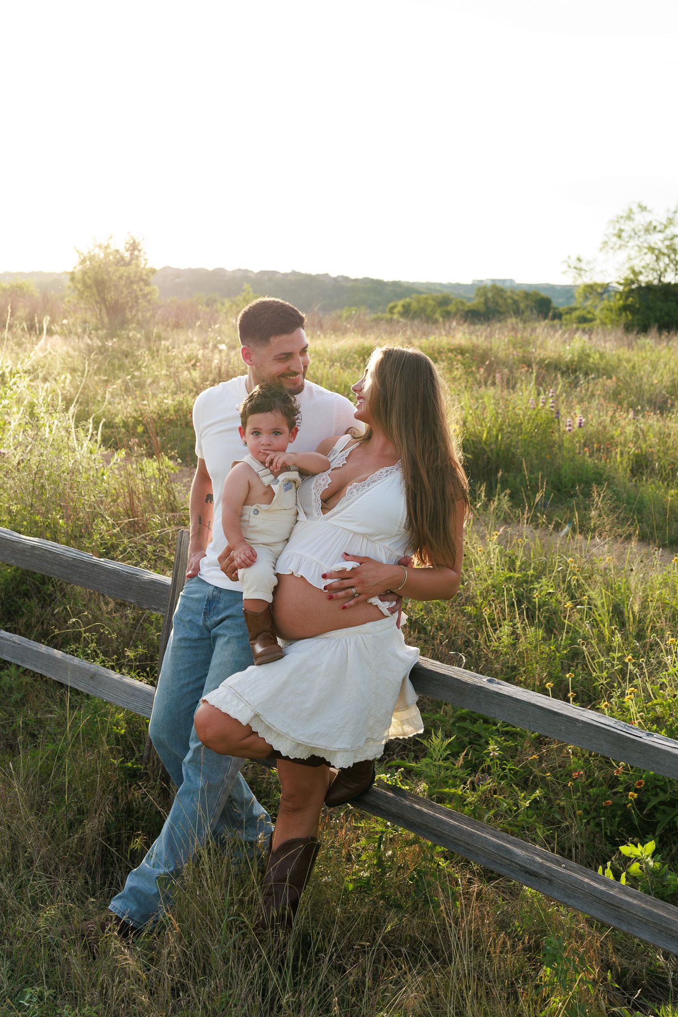 Pregnant woman in white dress with husband and toddler posing by rustic fence in sunlit field.