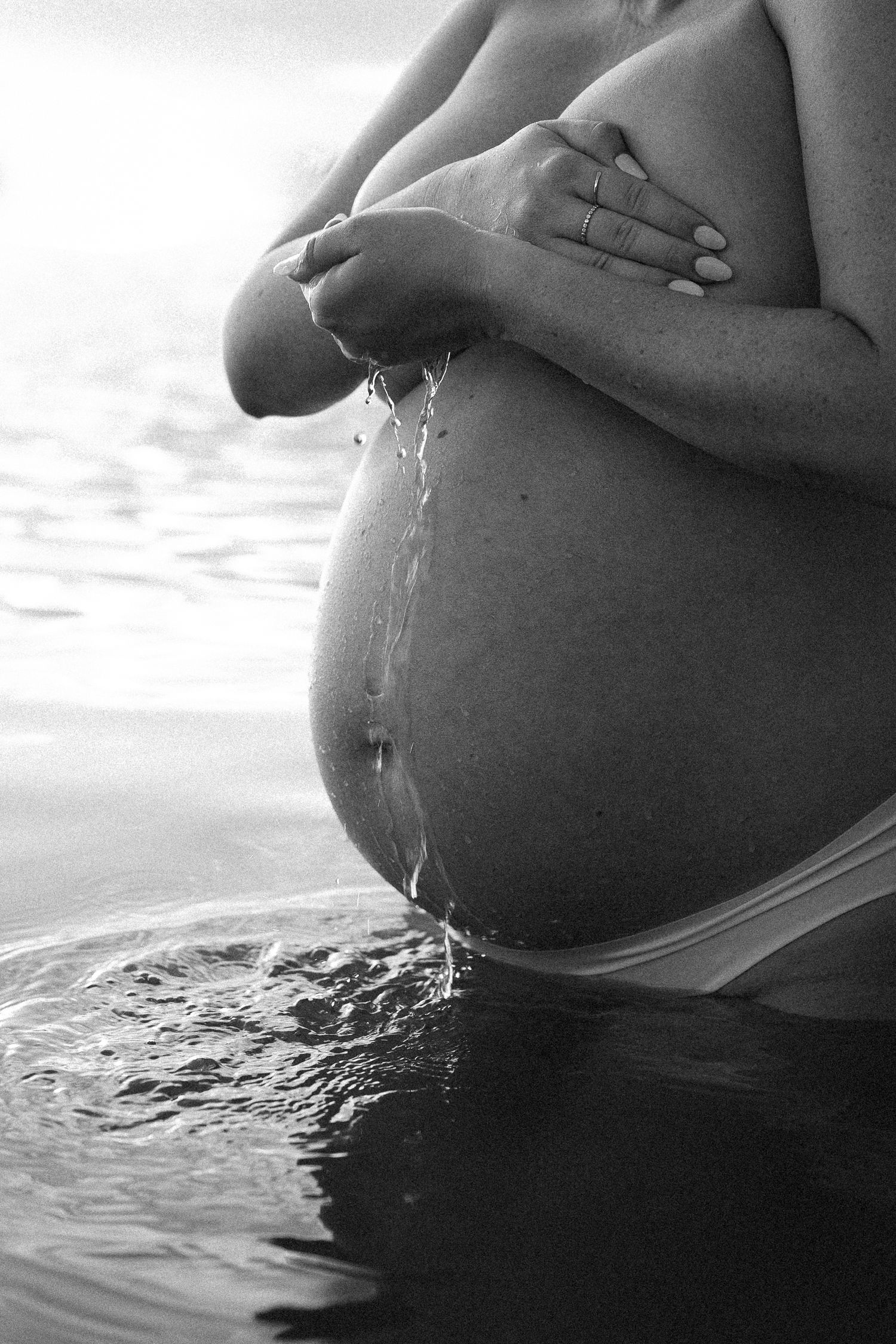 A black and white artistic sequence shows a pregnant belly in silhouette touching water, creating rippling reflections.