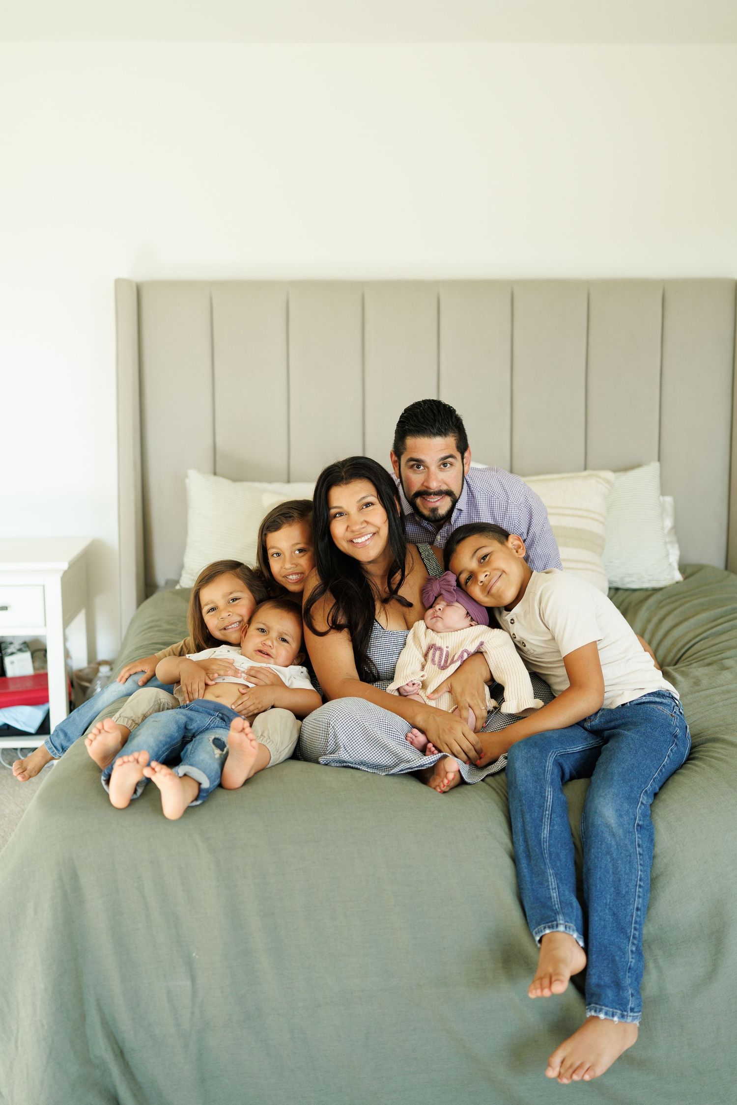 Happy family of six posing together on a bed, including parents and four children, one being a newborn baby.