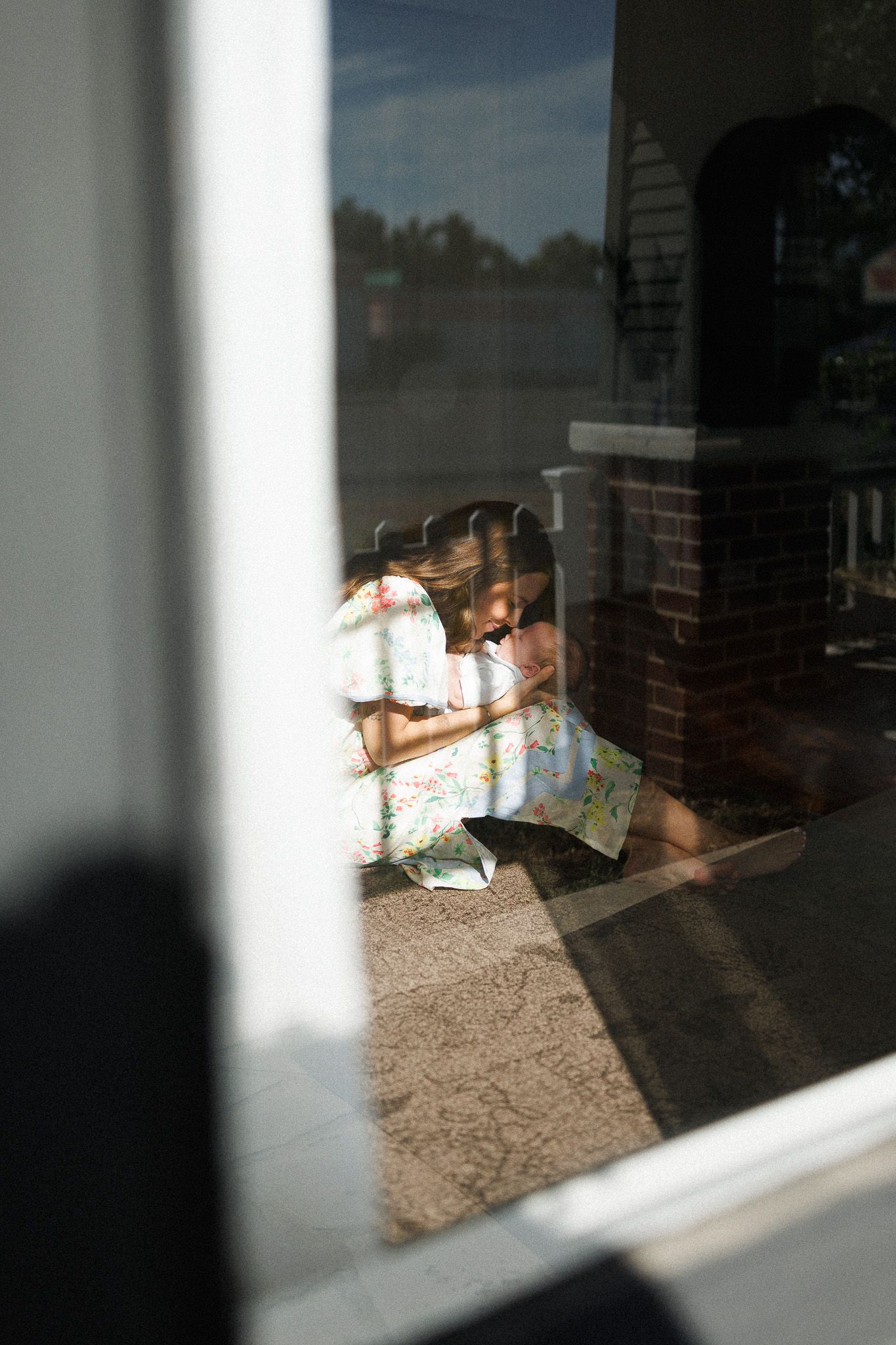 Young girl in floral dress reading a book on a sunlit porch, photographed through a glass window.