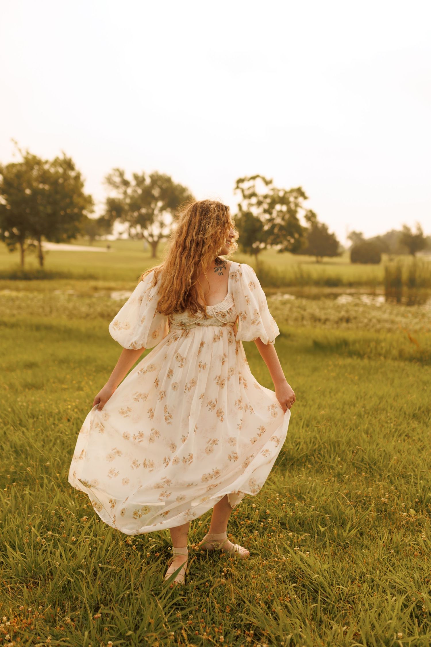 Woman in floral white dress twirls in a sunlit grassy field during golden hour at sunset.