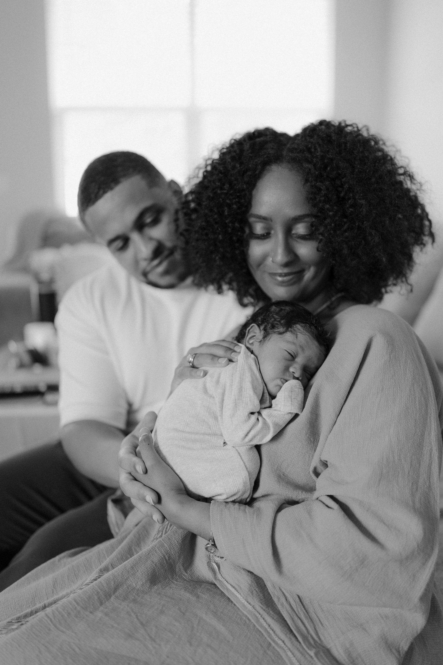 Black and white photo of parents lovingly cradling their sleeping newborn baby at home.