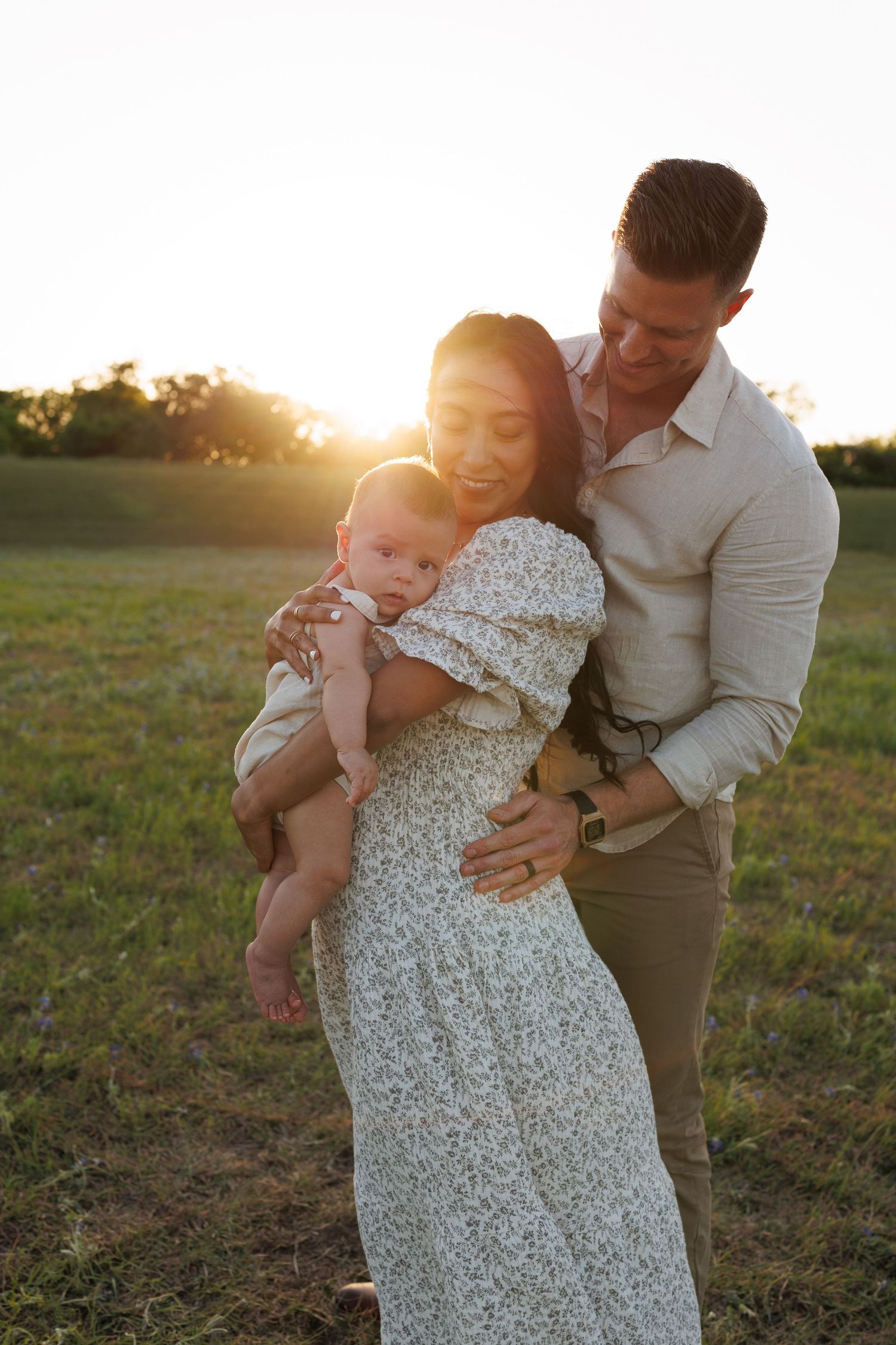 Family embracing together in a field at sunset, wearing elegant summer clothing as warm golden light glows behind them.