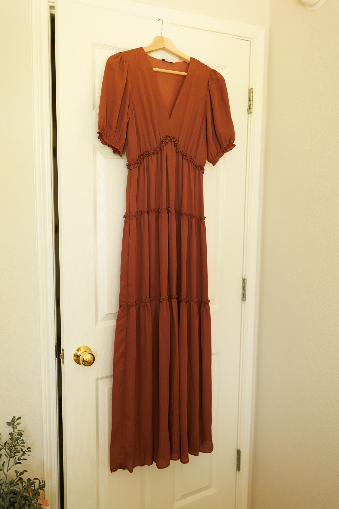 A rust-colored tiered babydoll dress with short sleeves hanging on a wooden hanger against a white door.