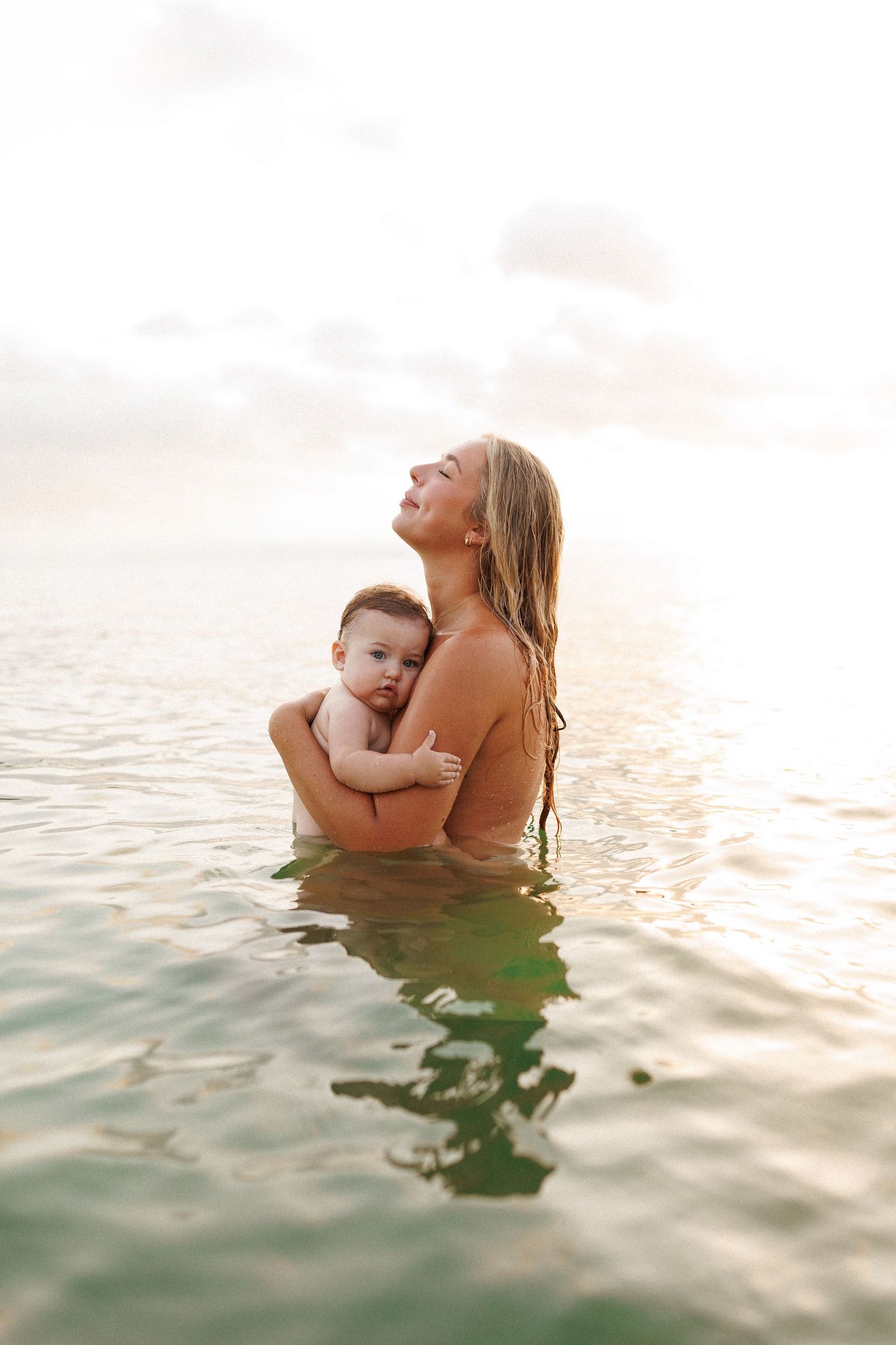 Two people embrace while standing in gentle ocean waters during sunset, creating a serene silhouette against the sky.