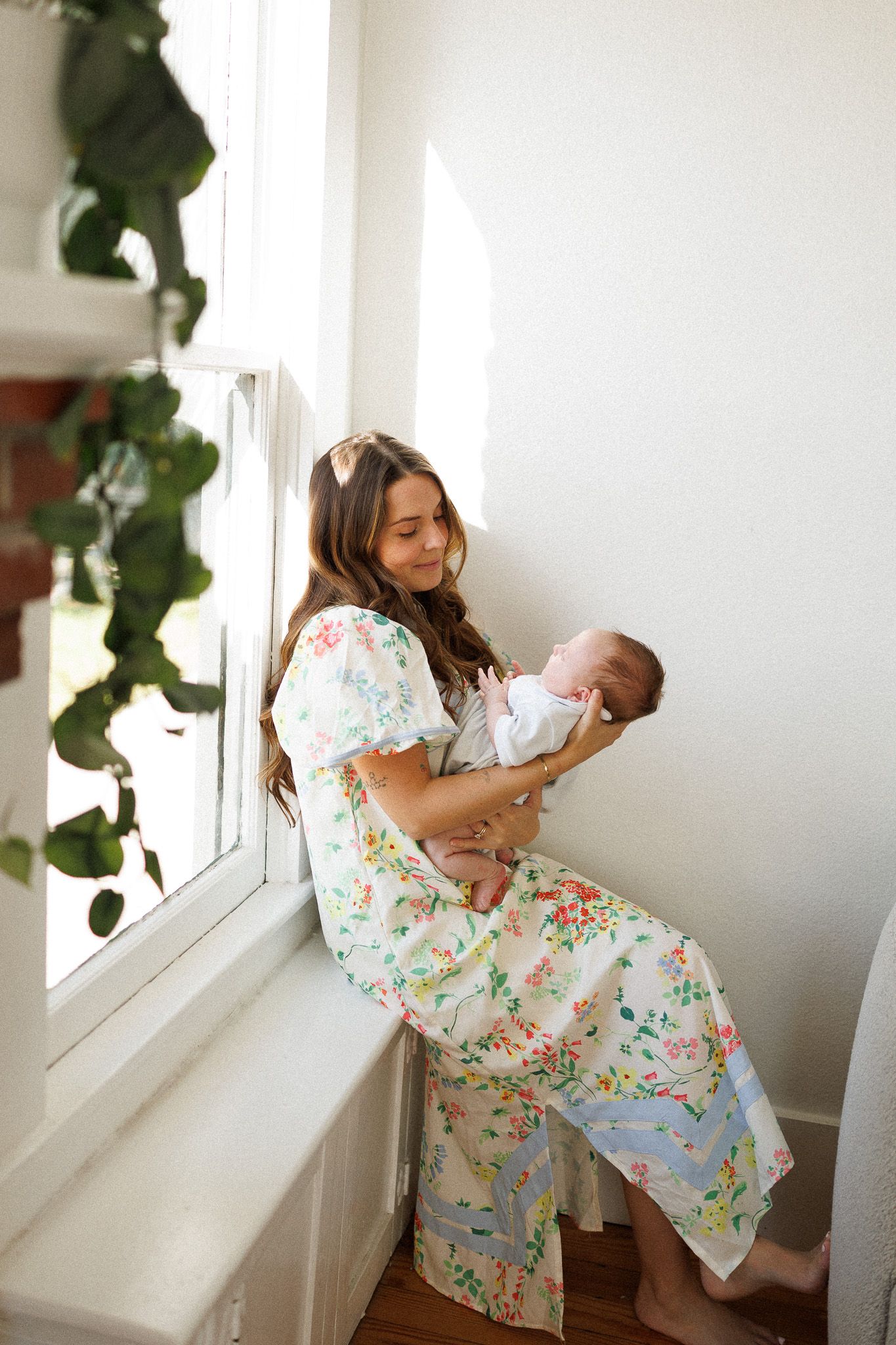 Mother in floral dress tenderly holds sleeping newborn baby while sitting by a bright window.