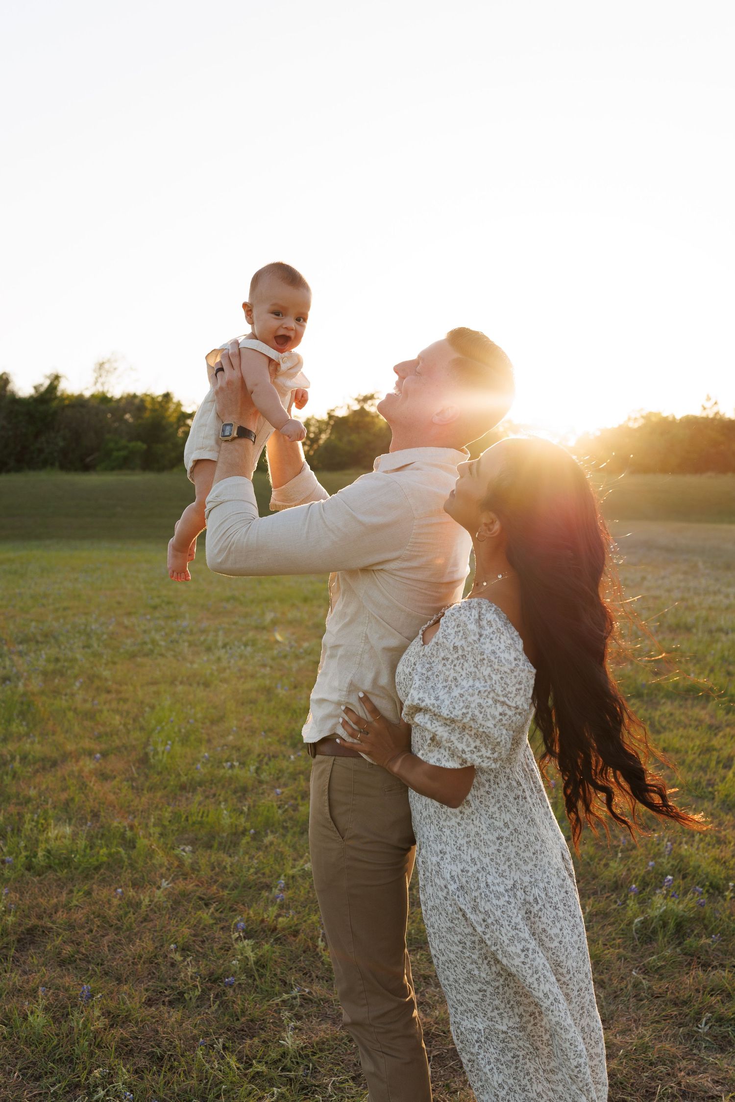 A couple shares a joyful moment with their baby in a sunlit field during golden hour.