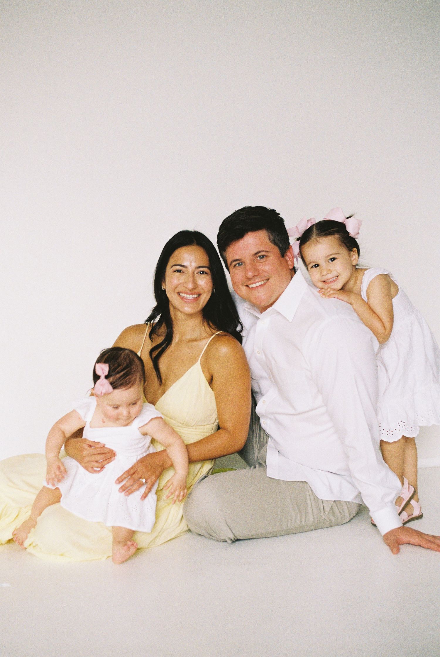 A smiling family of four poses together on a white studio floor, with mom in yellow, dad in white, and two young daughters.