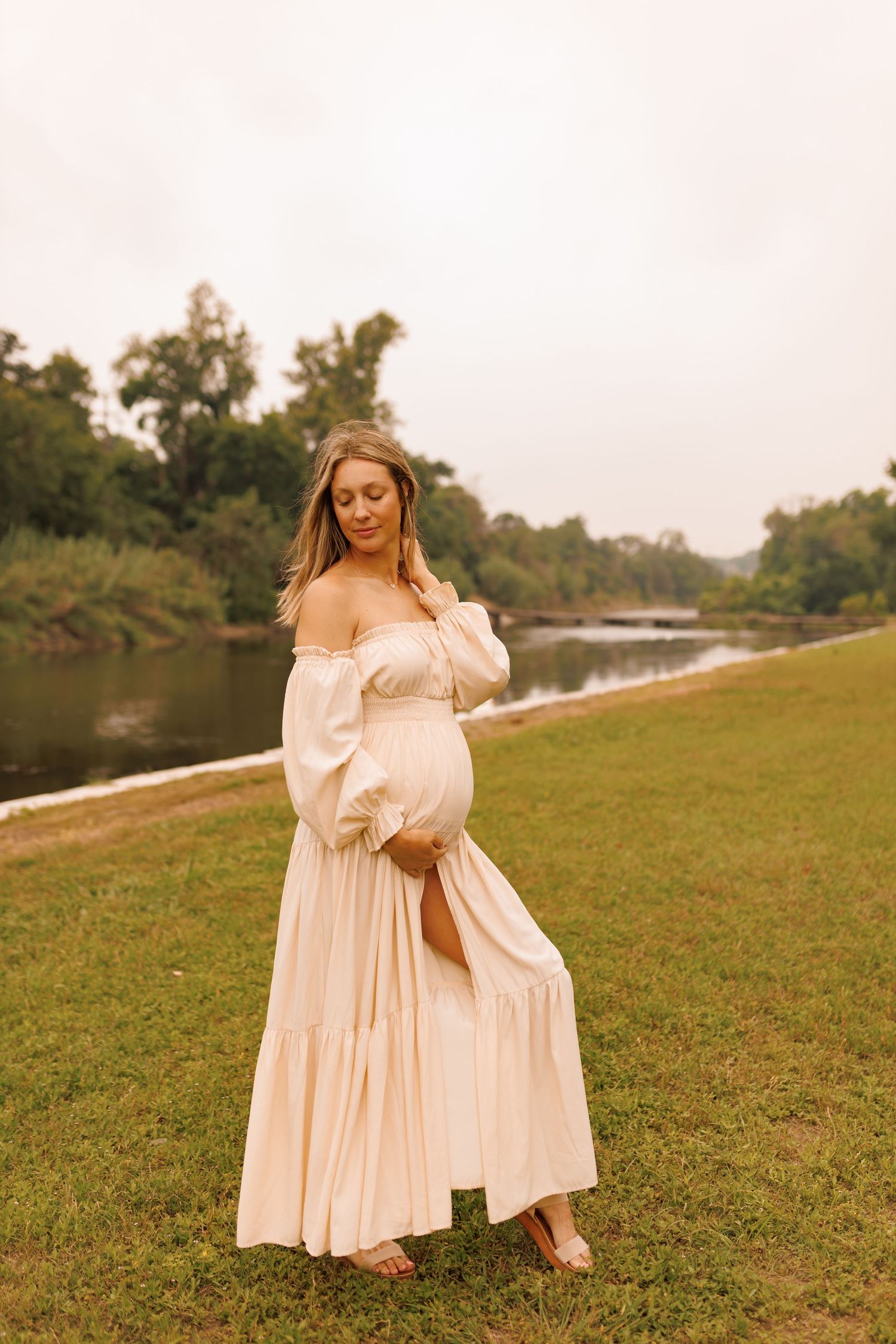 A person in a flowy cream-colored dress poses gracefully on a grassy riverbank during golden hour.