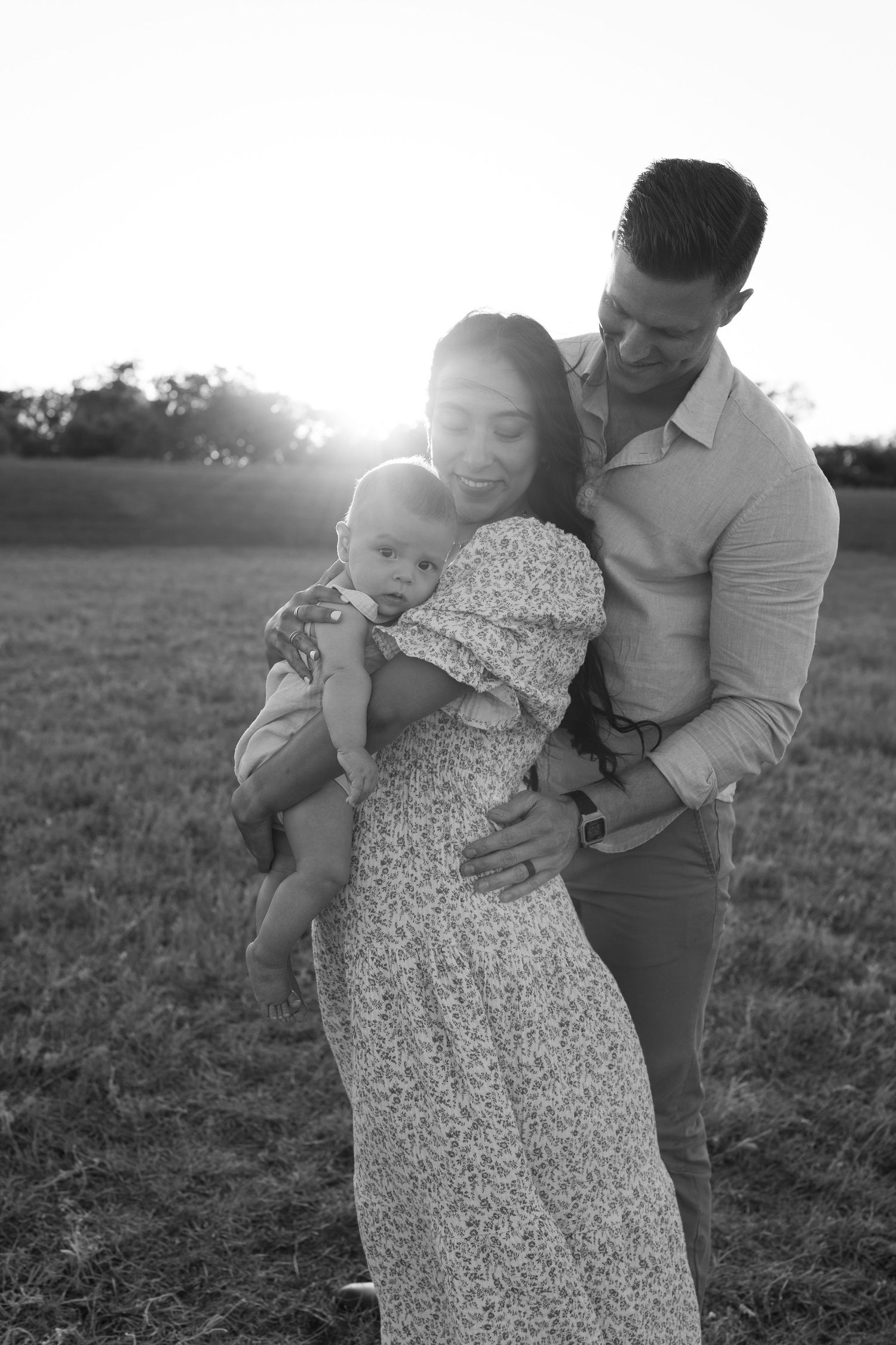 A family embraces in a sunny field, wearing casual clothes in a tender black and white portrait moment.