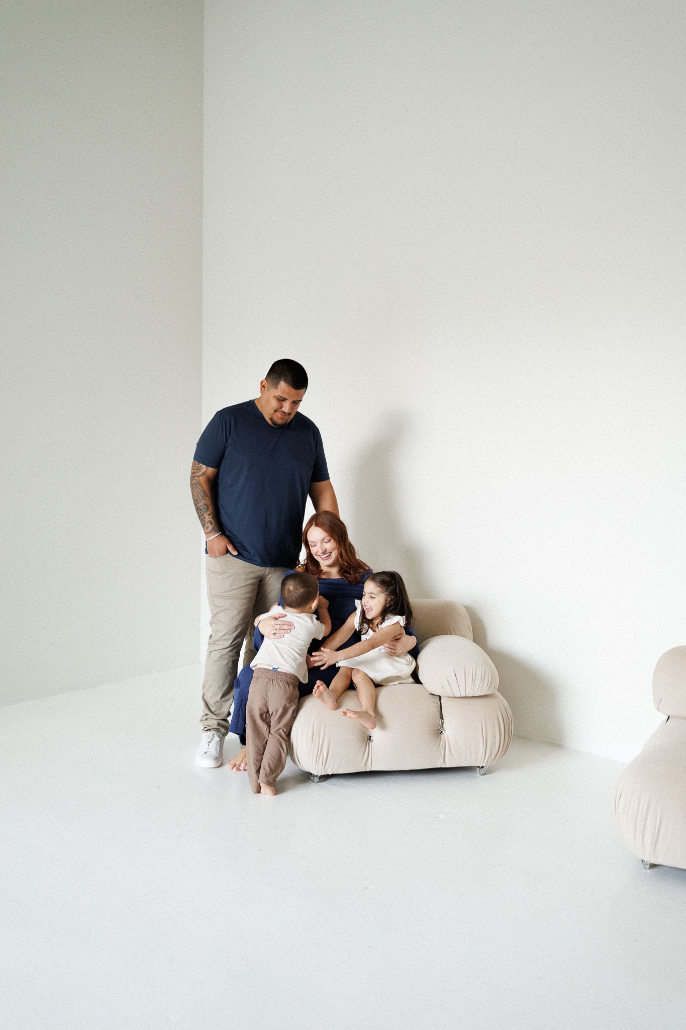 Family members share tender moments together while relaxing on a cream-colored couch in a minimalist white room.