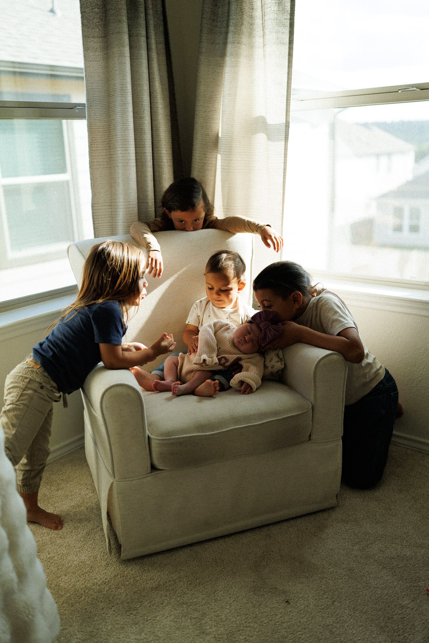 Children gathered around a white armchair admiring a newborn baby in a bright living room.