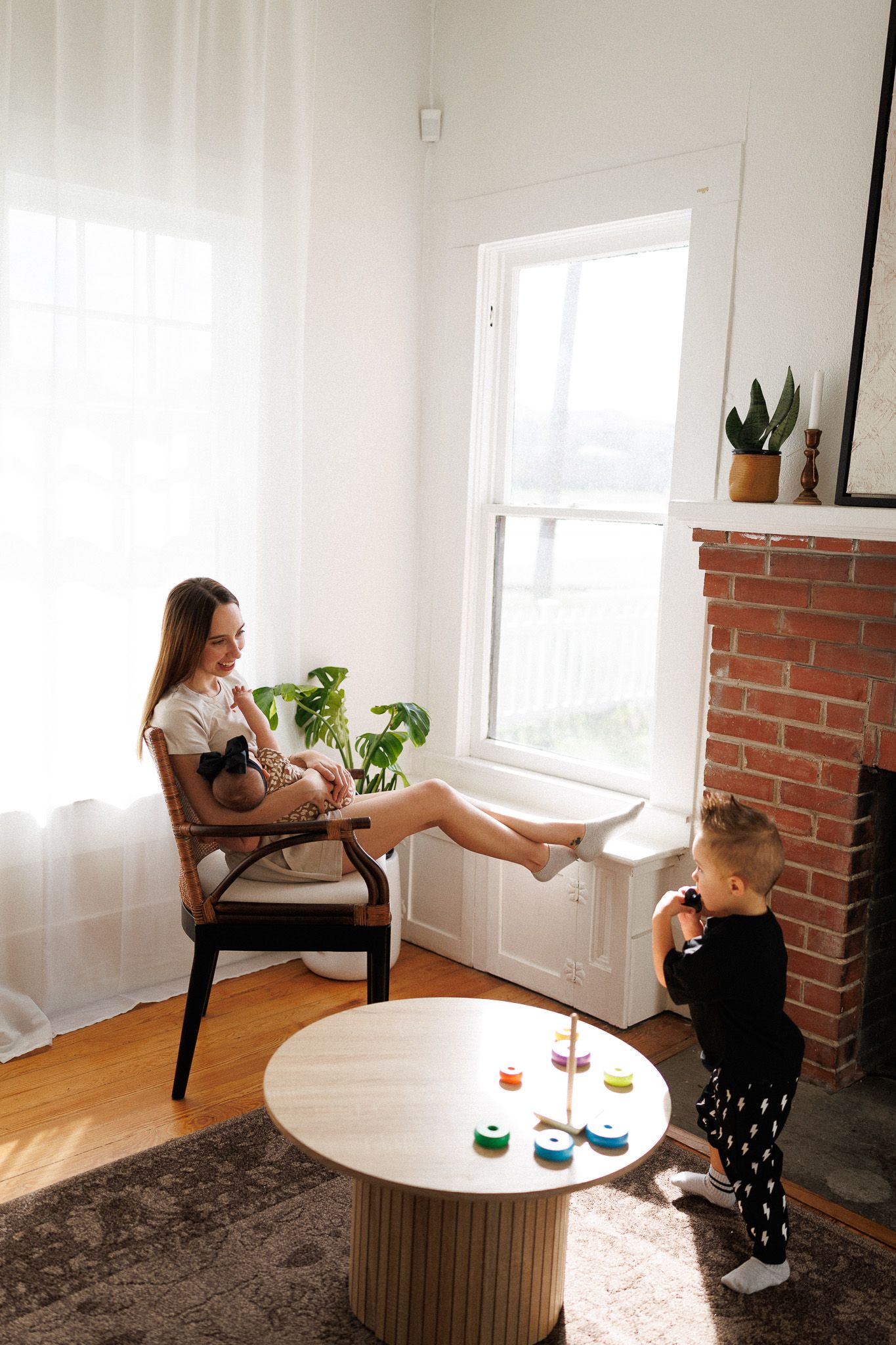 A woman sits in a chair holding a potted plant, with a young child playing on the floor in a cozy, sunlit room.