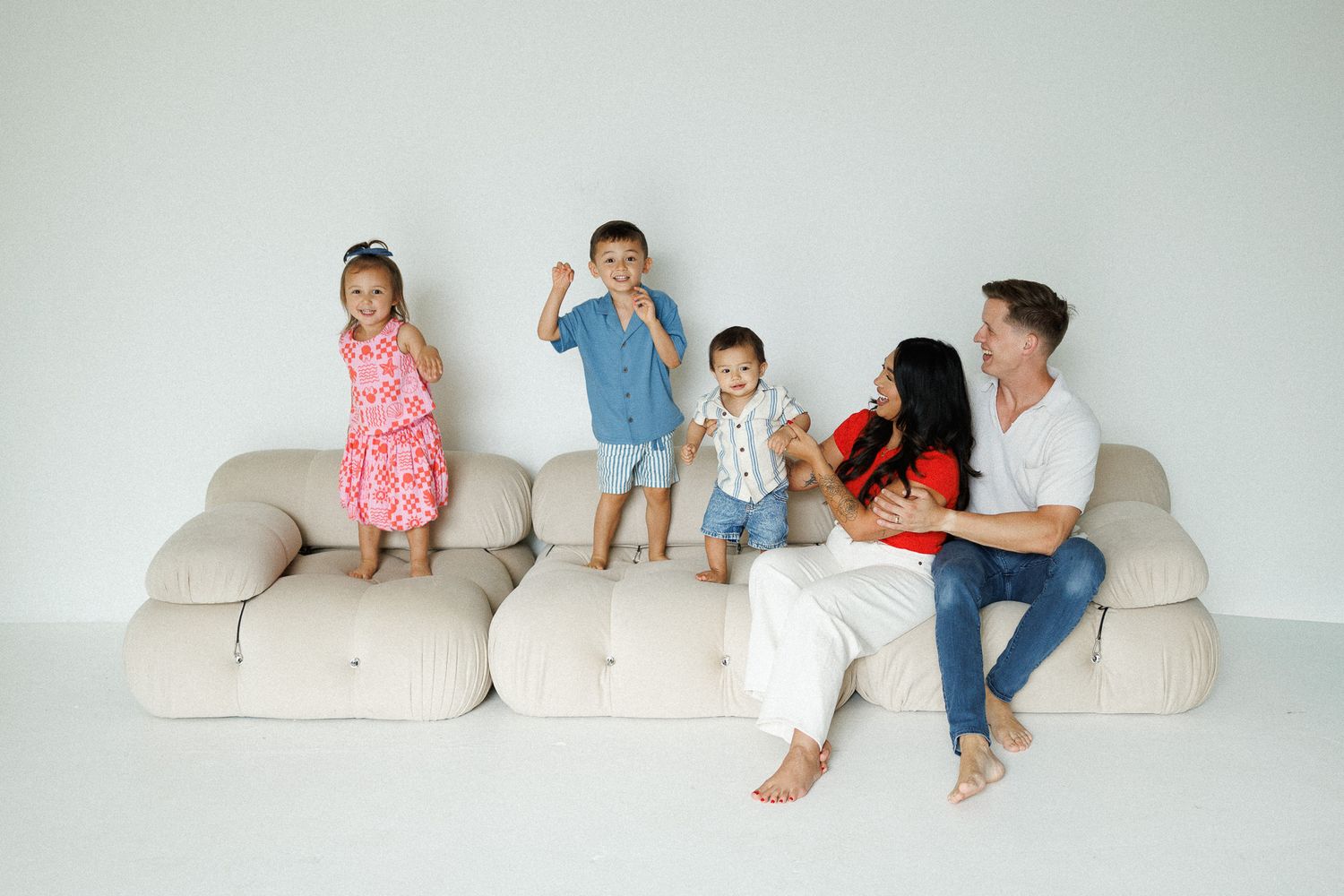 Happy family with three young children playing on a large beige sectional sofa against a white background.