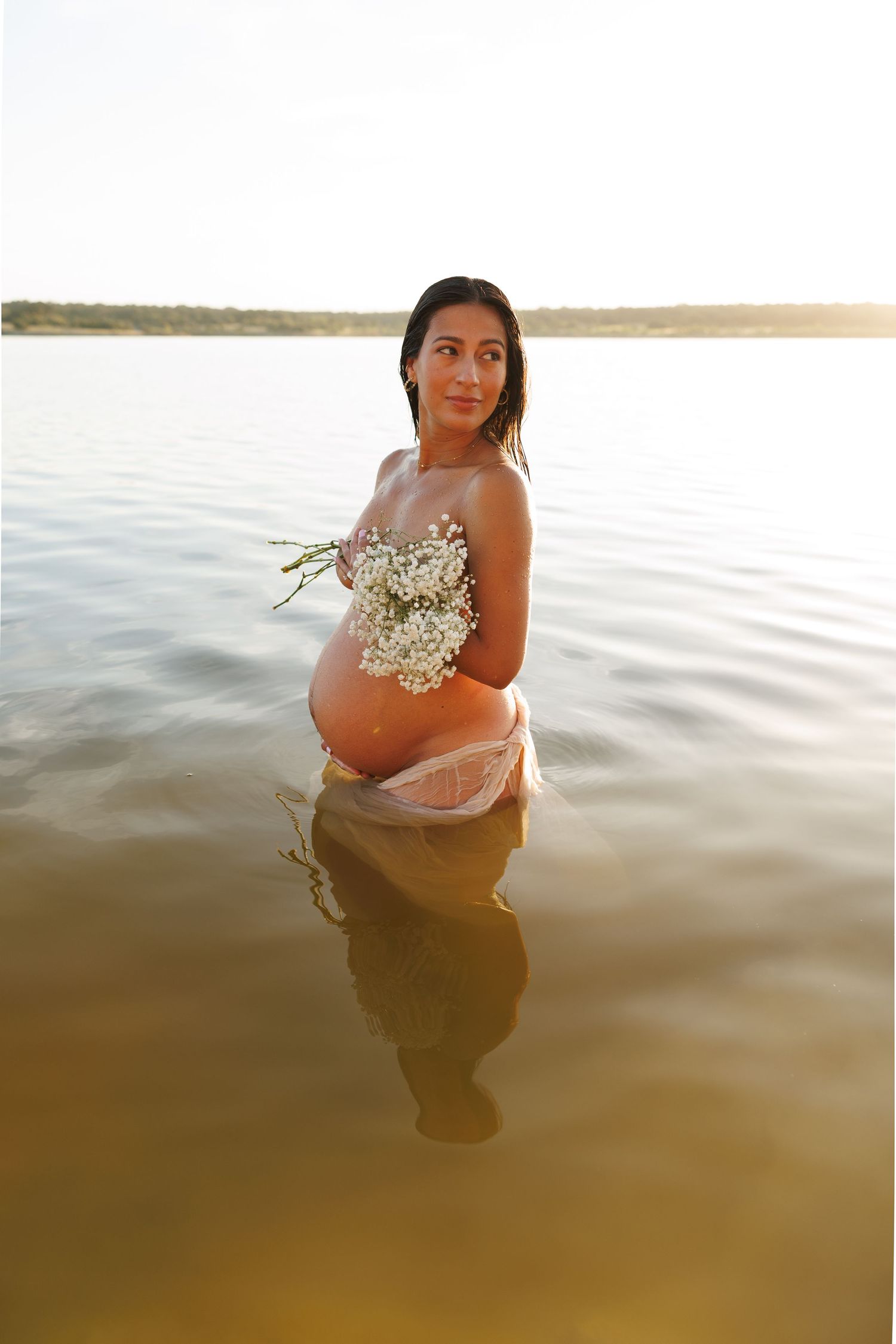 An expectant mother wearing a white floral top stands in calm lake waters at sunset creating a beautiful reflection.