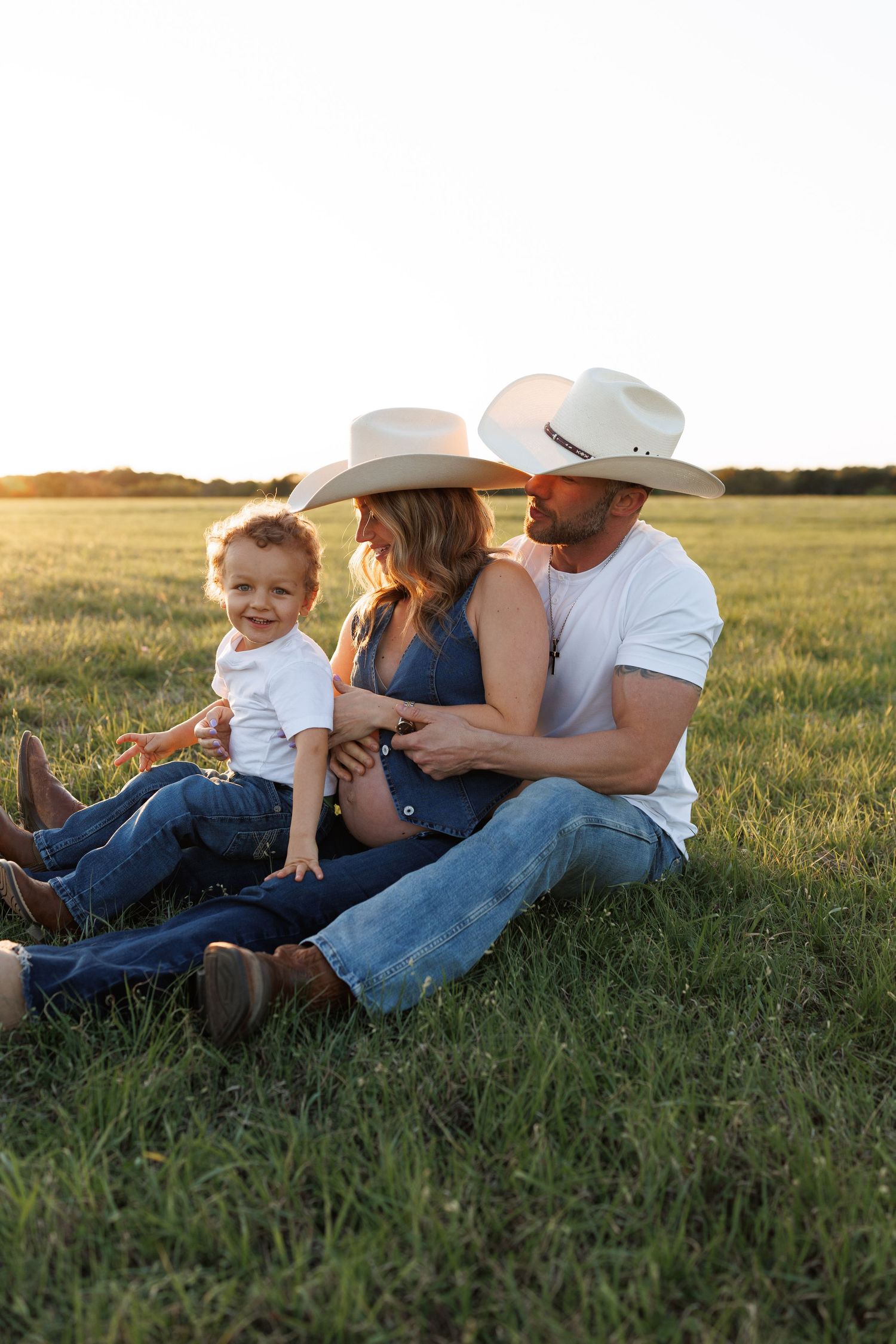 Three people wearing cowboy hats sit together in a grassy field at sunset, creating a cozy country scene.