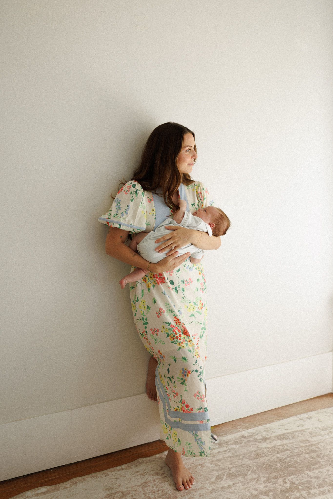 Woman in floral maxi dress holding newborn baby against white wall, barefoot on hardwood floor.