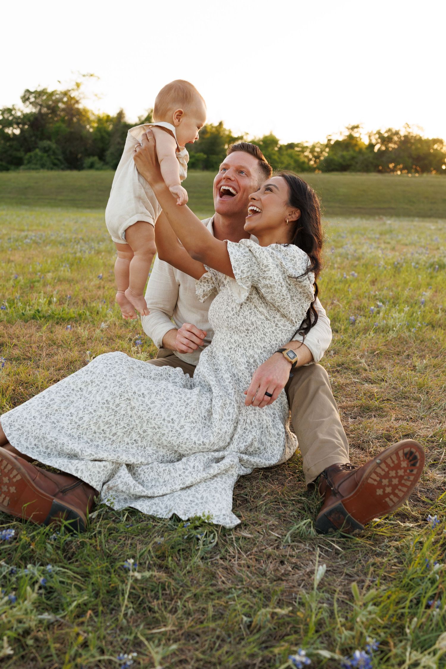 Family laughs joyfully together while sitting in a grassy field at sunset wearing rustic countryside attire.