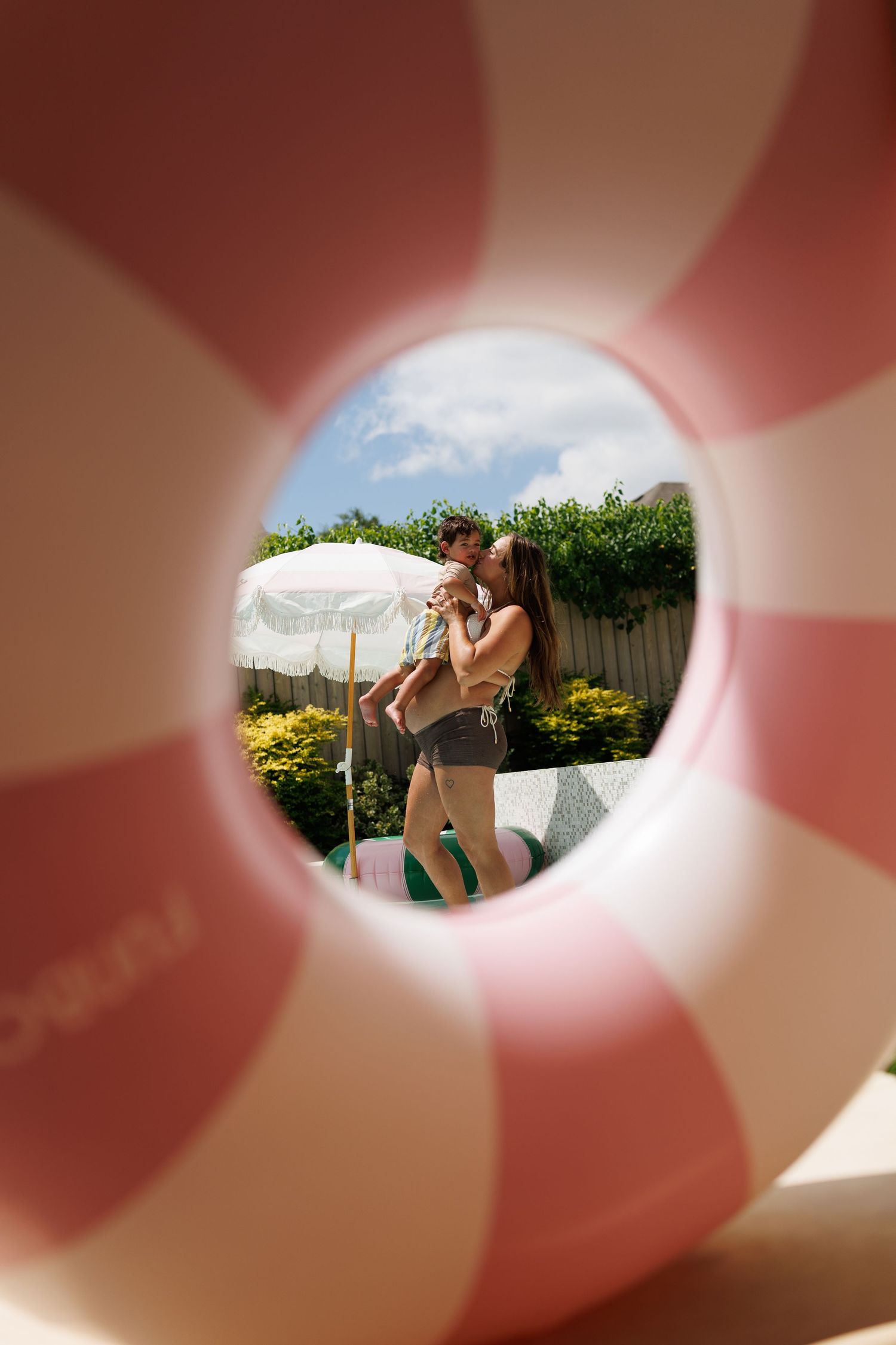 A couple kisses while holding a baby, viewed through a pink and white inflatable pool ring on a sunny day.