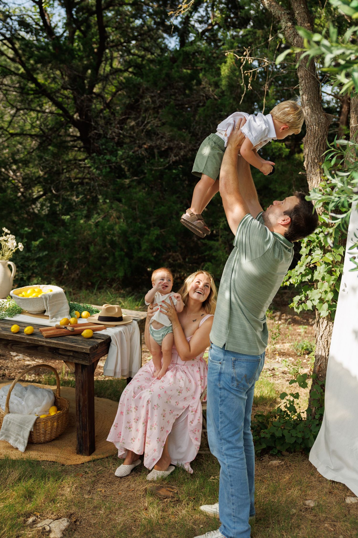 A joyful family outdoors, dad lifting toddler high, mom holding baby, beside a rustic table with lemons in a sunny garden.