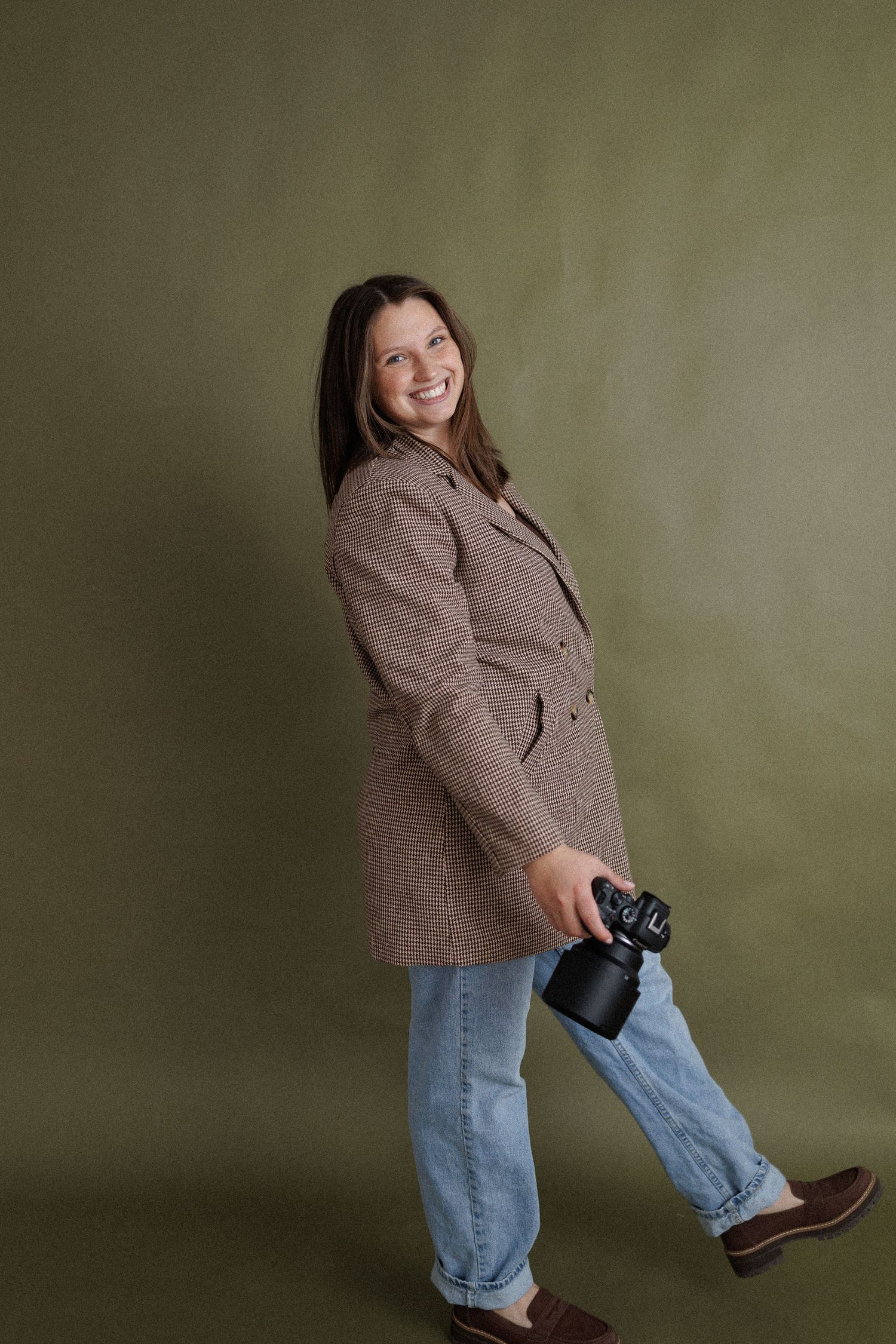 A photographer wearing a brown tweed blazer and jeans poses playfully with a camera against an olive green background.