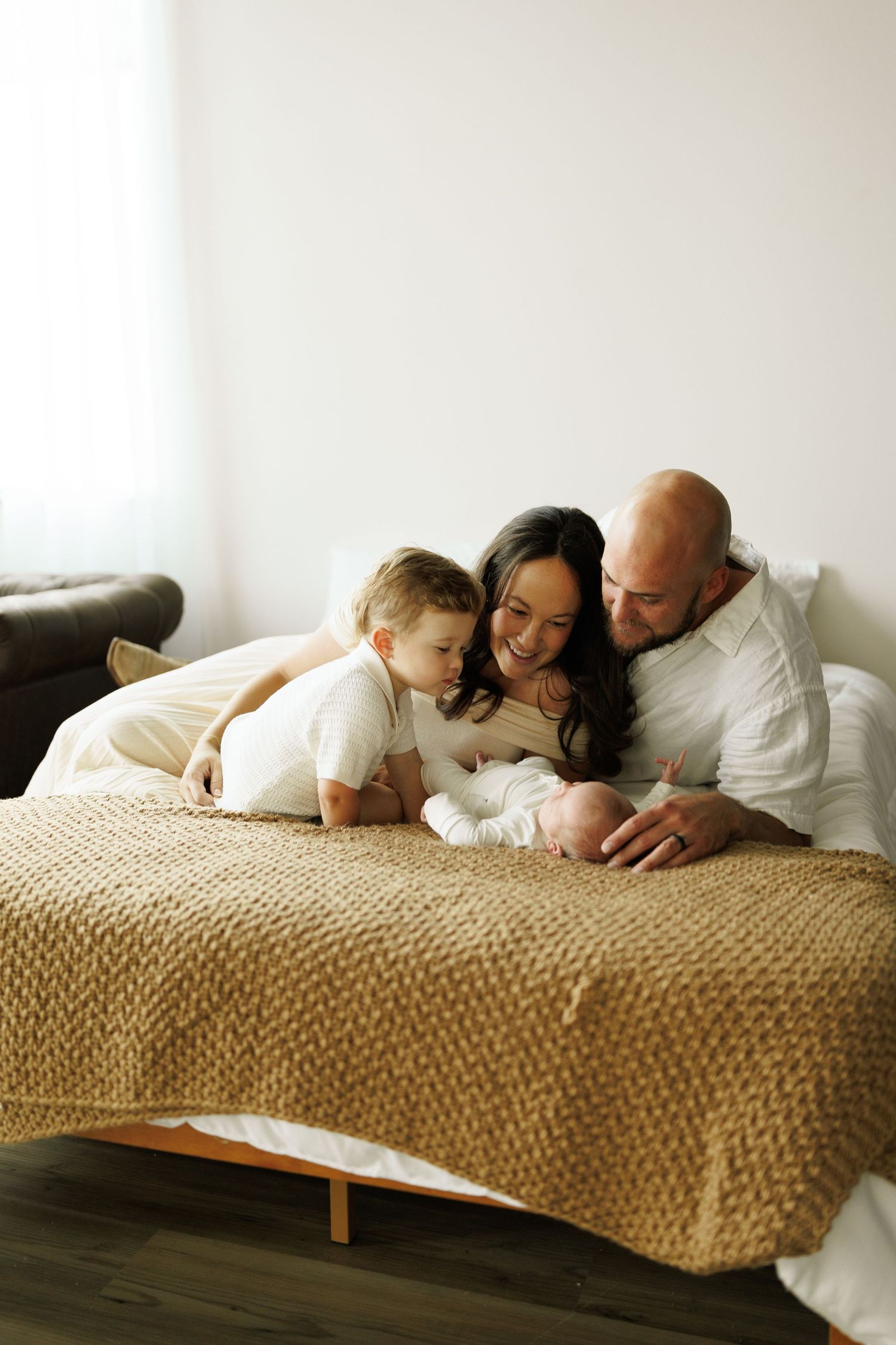 A family of three cuddles together on a bed with a knit beige blanket in a bright minimalist room.