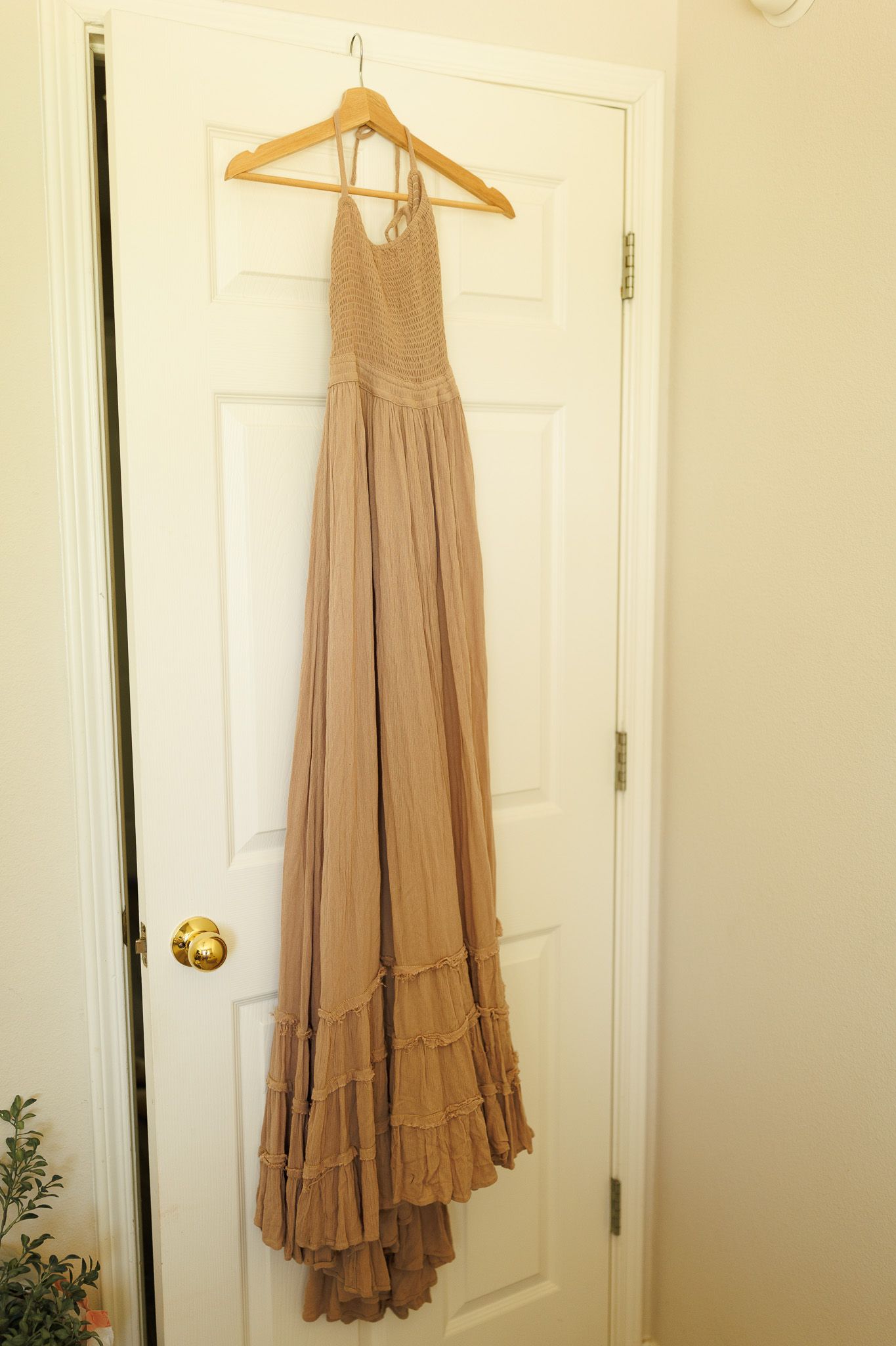 A beige maxi dress with ruffled hem hangs on a wooden hanger against a white door.