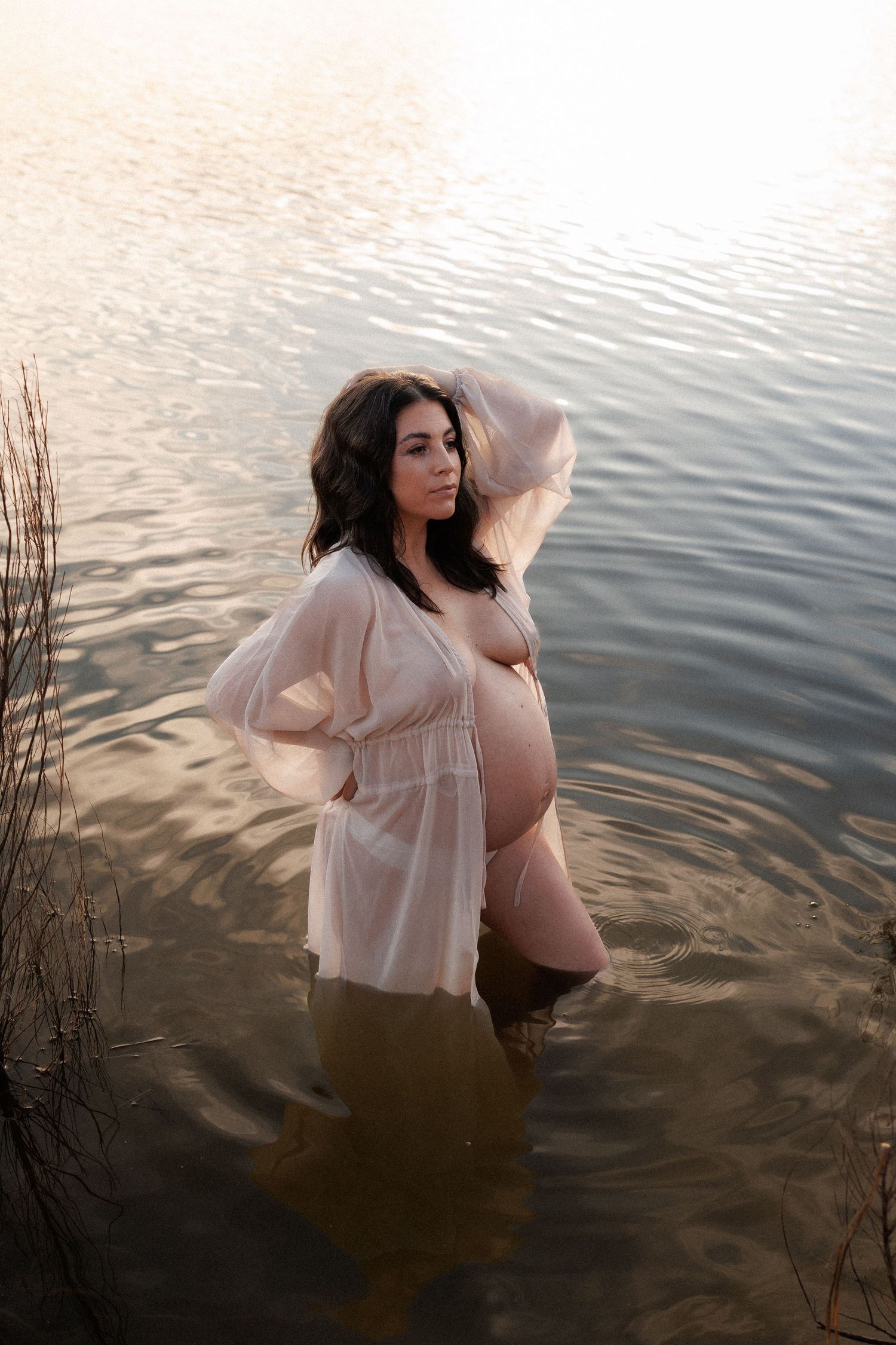 A pregnant woman in sheer white dress stands in shallow water during golden sunset light, capturing a serene maternity moment.