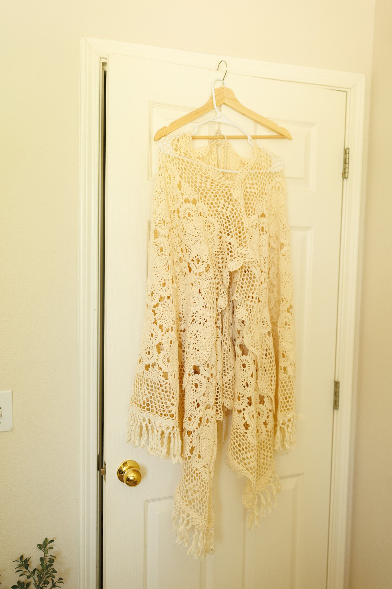 A delicate cream-colored crochet shawl with intricate lace patterns hangs on a wooden hanger against a white door.