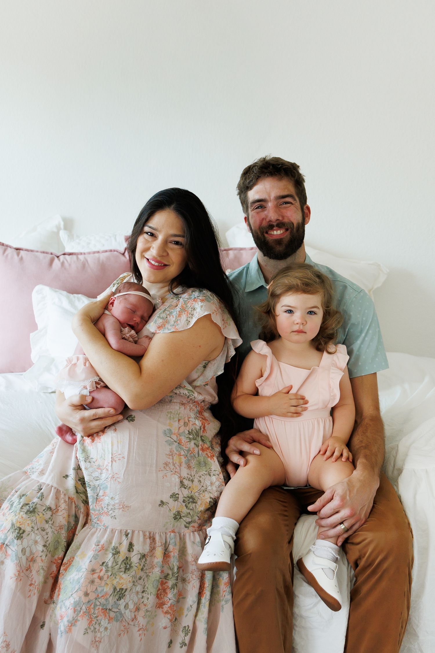 Happy family of four posing on a white bed, mother holding newborn baby, father with toddler girl in pink romper.