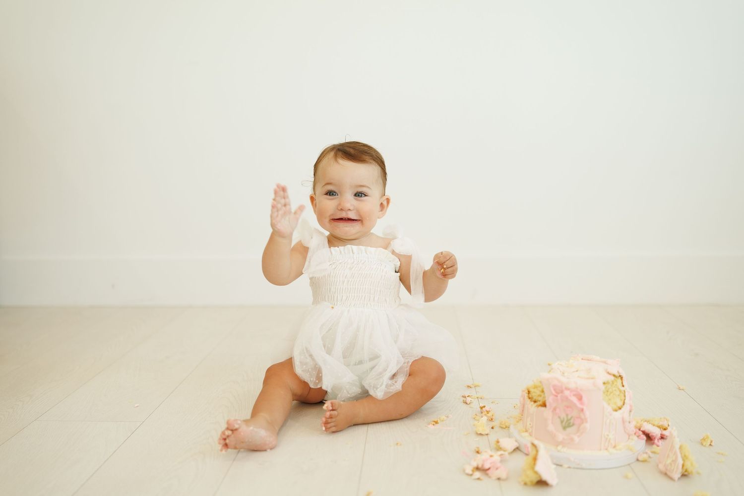 Baby wearing white dress sitting on light wooden floor with pink floral decorations nearby.