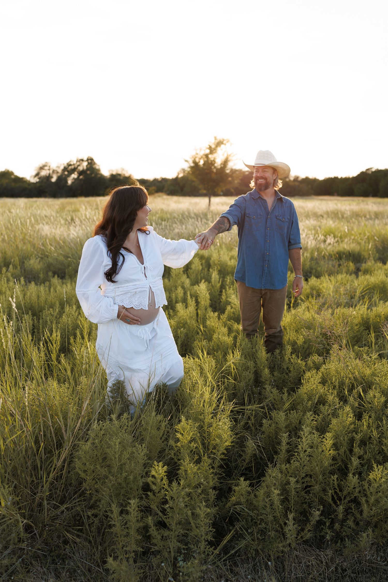 A couple walks together through a sunlit field of tall grass during golden hour creating a romantic silhouette.