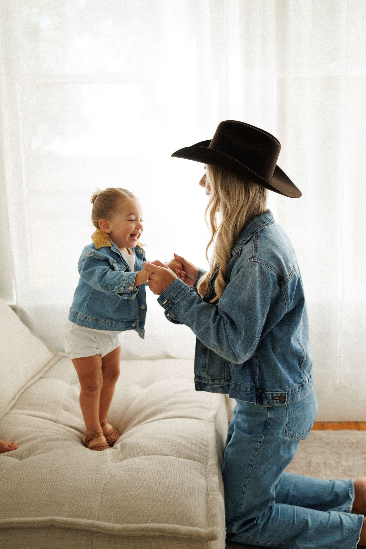 A woman in denim and black cowboy hat plays with a laughing toddler girl in matching denim jacket on a white sofa.
