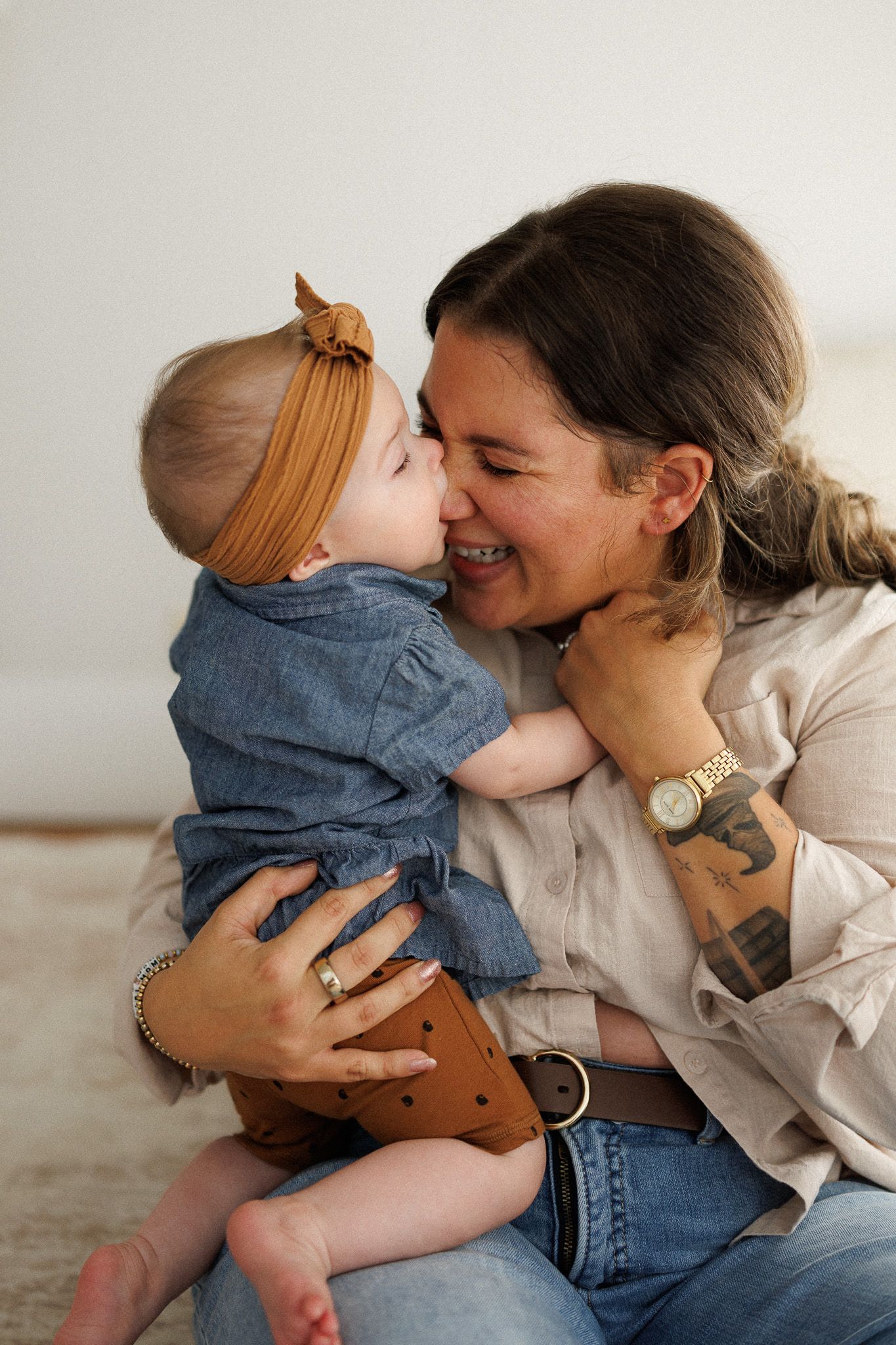 Tattooed mother smiling as baby in denim outfit and bow headband nuzzles her cheek.