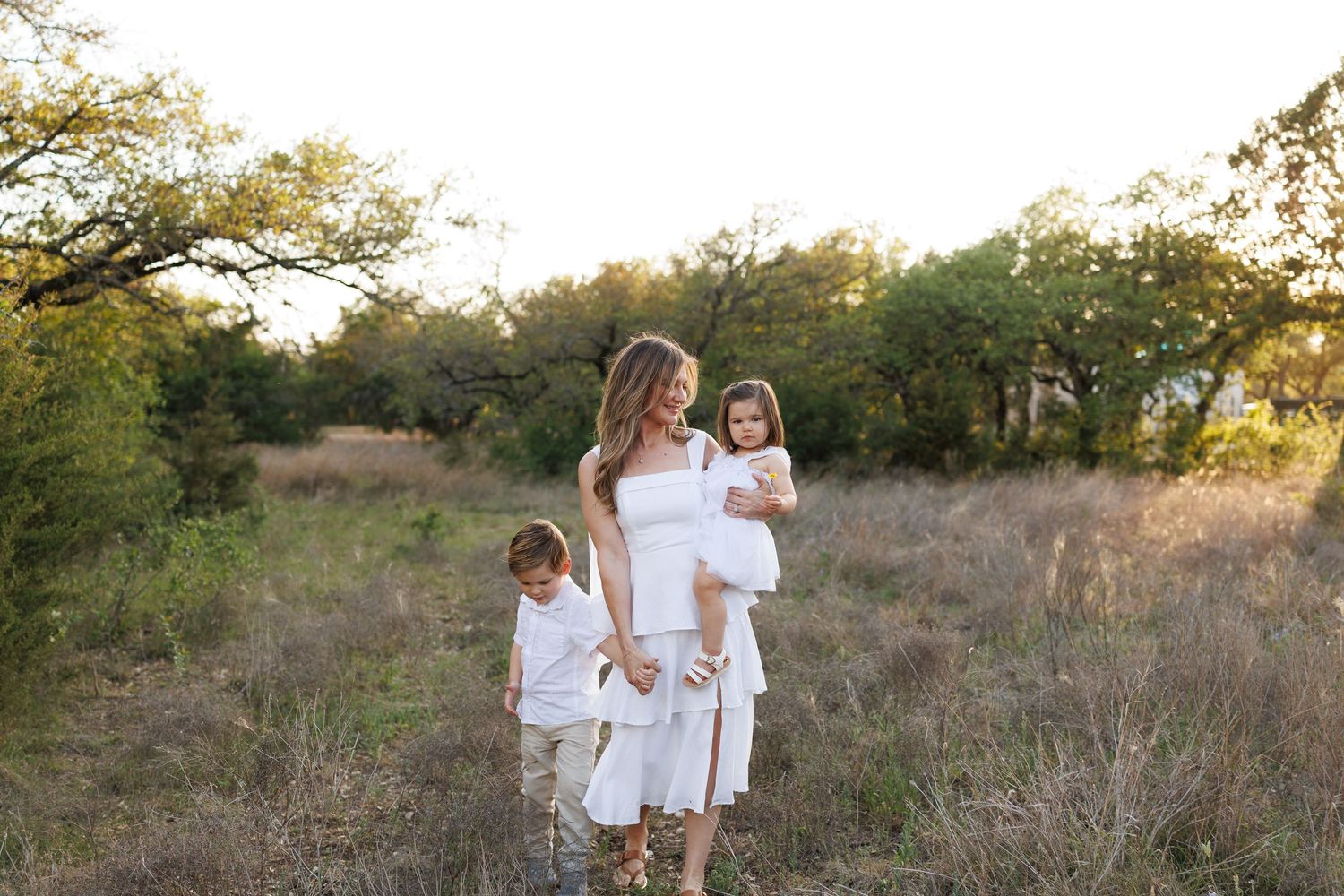 A mother in a white dress holds her daughter while her son stands beside her in a sunlit meadow.