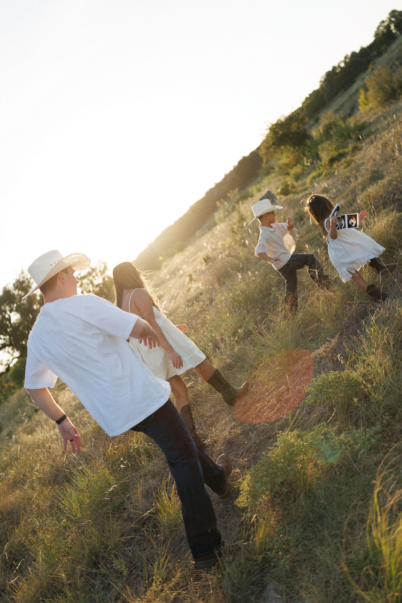 Family in white outfits on a sunny hillside, with children holding an ultrasound photo during a pregnancy announcement.