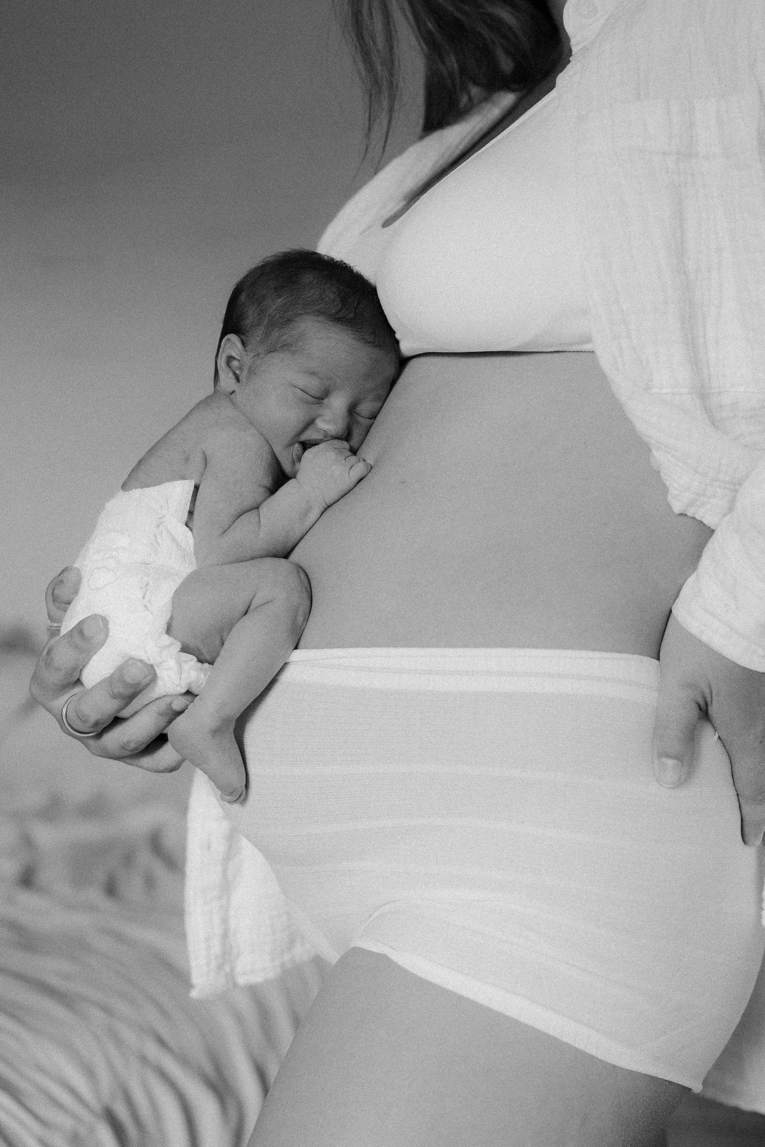 Black and white photo of a pregnant woman holding a sleeping newborn baby against her bare belly.