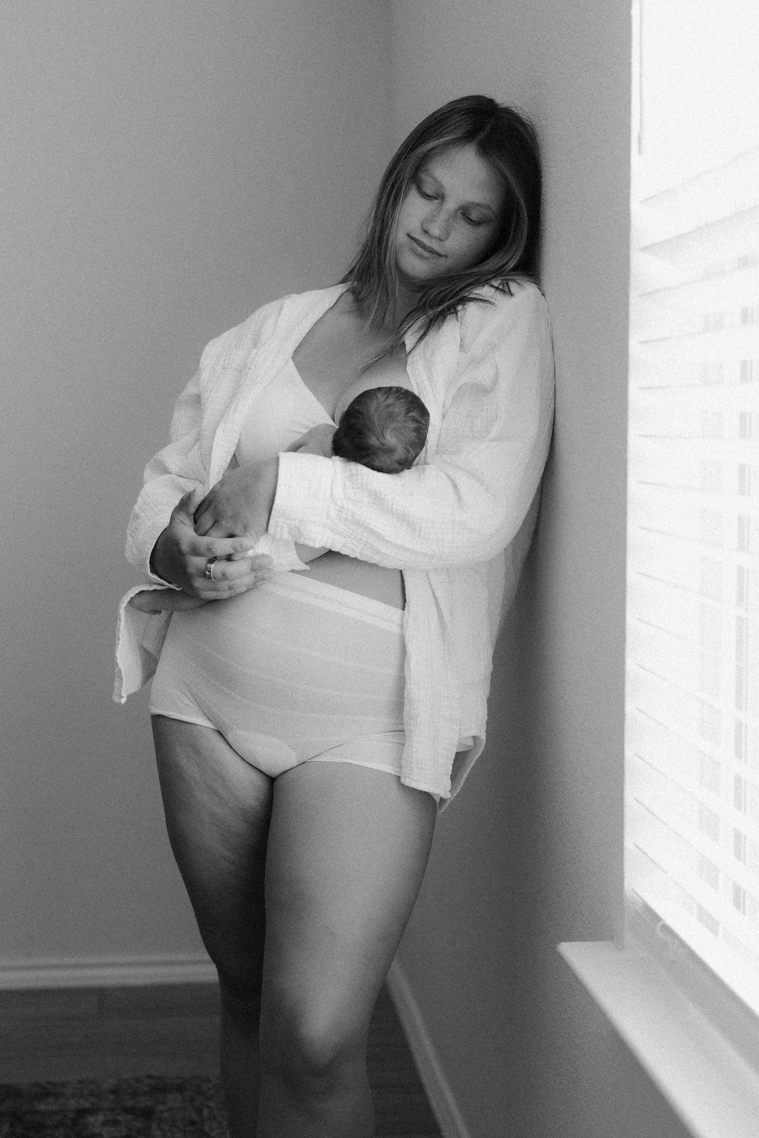 Black and white photo of a new mother in white robe holding her newborn baby while leaning against a wall.