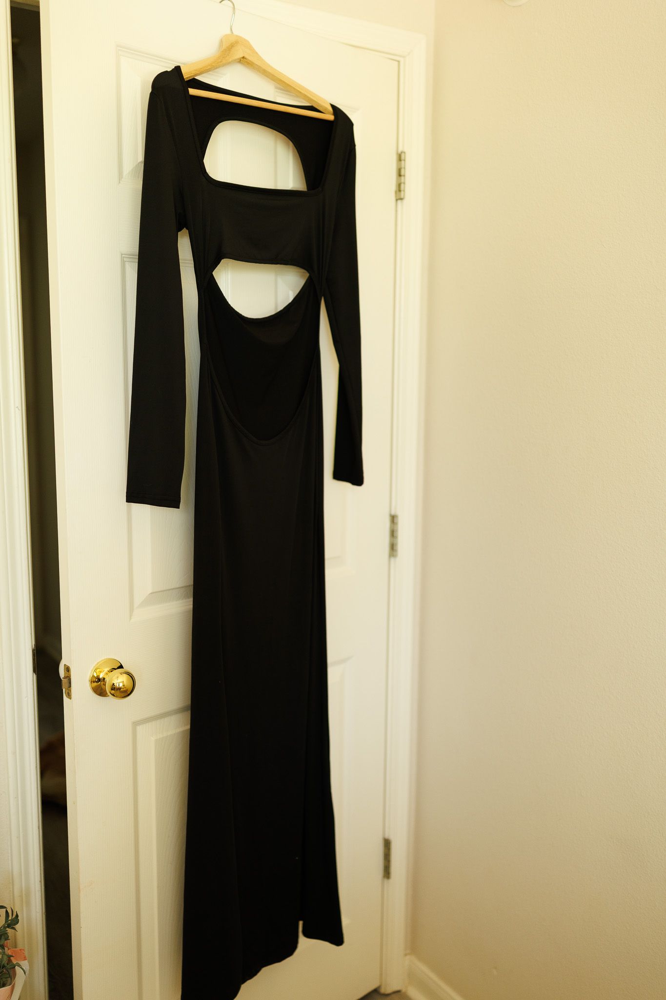 A black floor-length dress with long sleeves and cutout details hangs on a wooden hanger against a white door.