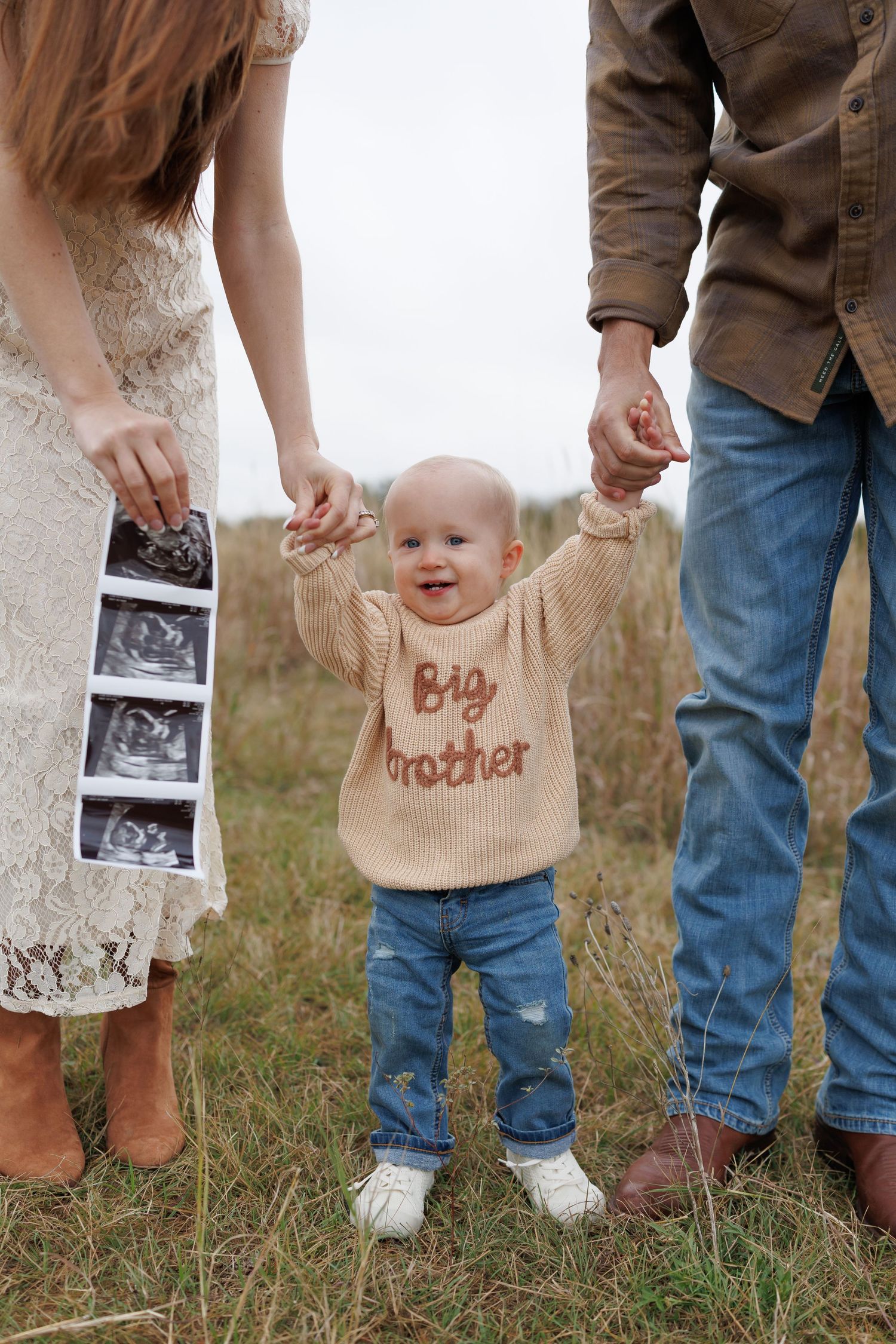 Pregnancy announcement photo with ultrasound strip and toddler wearing 'Big Brother' sweater holding parents' hands.