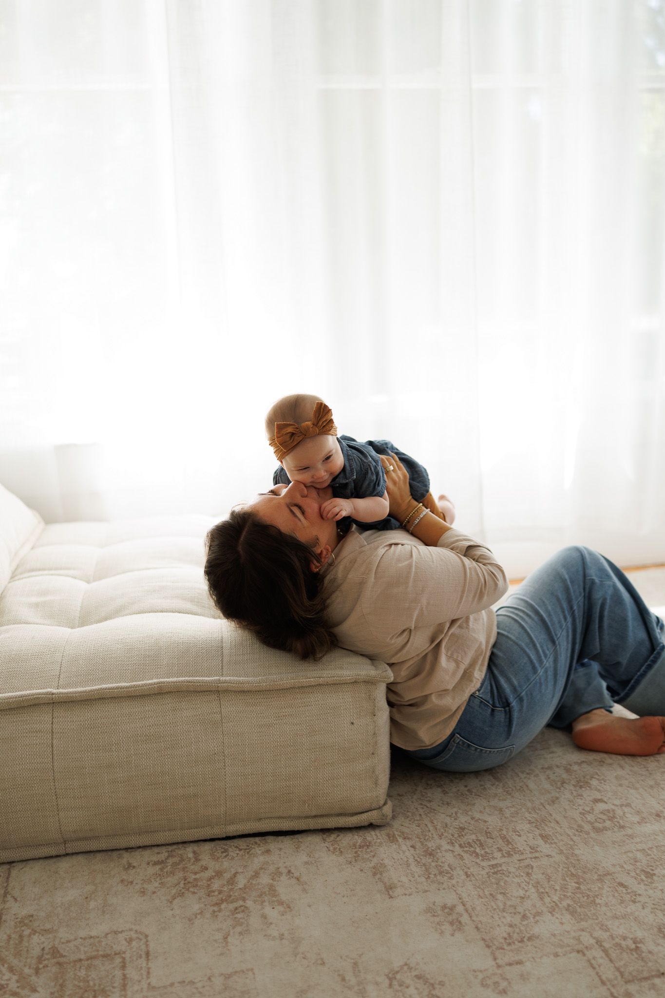 A mother lying on the floor lifts her smiling baby above her face near a white sofa in a bright living room.