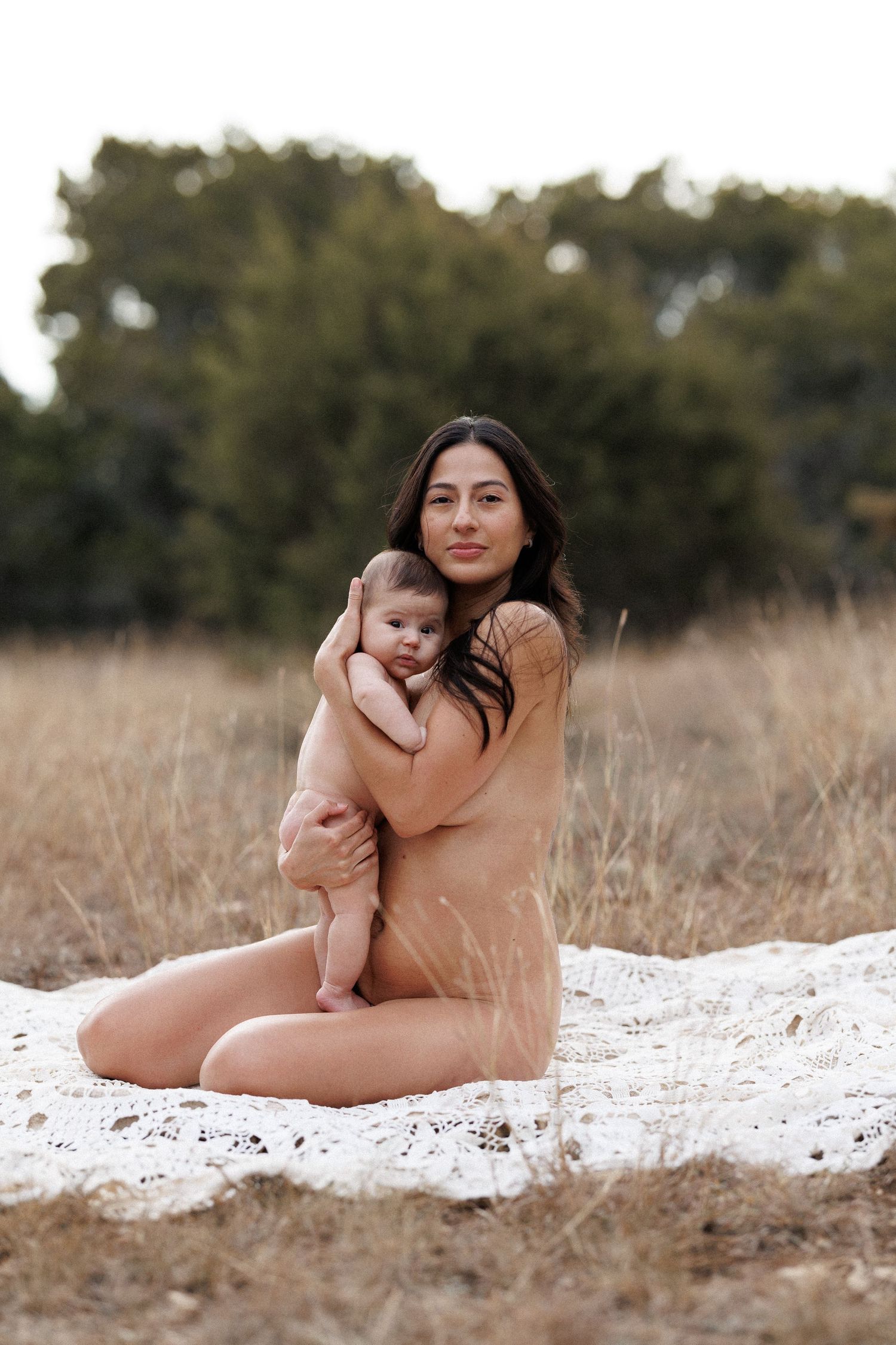 A mother and baby pose together on a white blanket in a rustic outdoor field setting during a photo session.