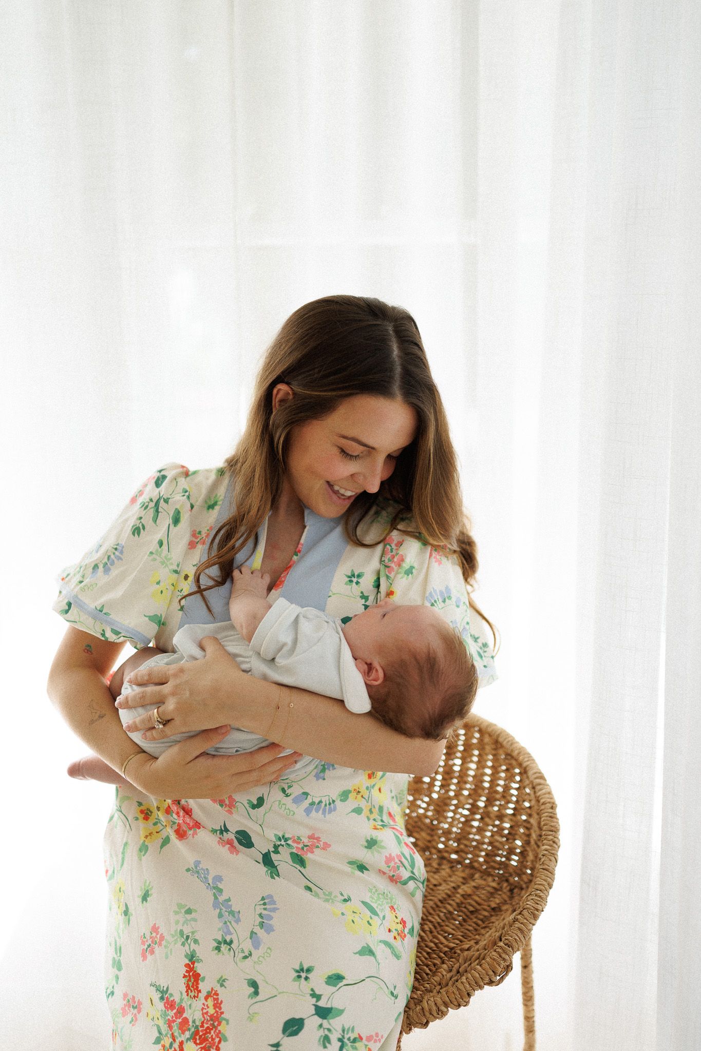 A smiling mother in a floral dress tenderly holds her newborn baby near a wicker bassinet by sheer white curtains.