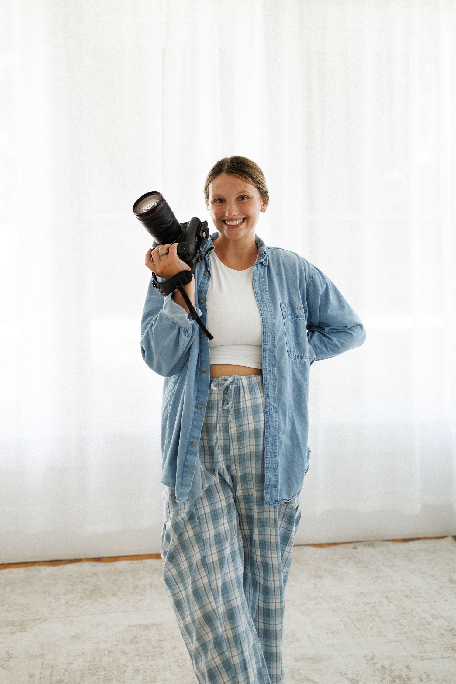 Smiling woman holding a DSLR camera on her shoulder, wearing a blue shirt and plaid pants, standing indoors.