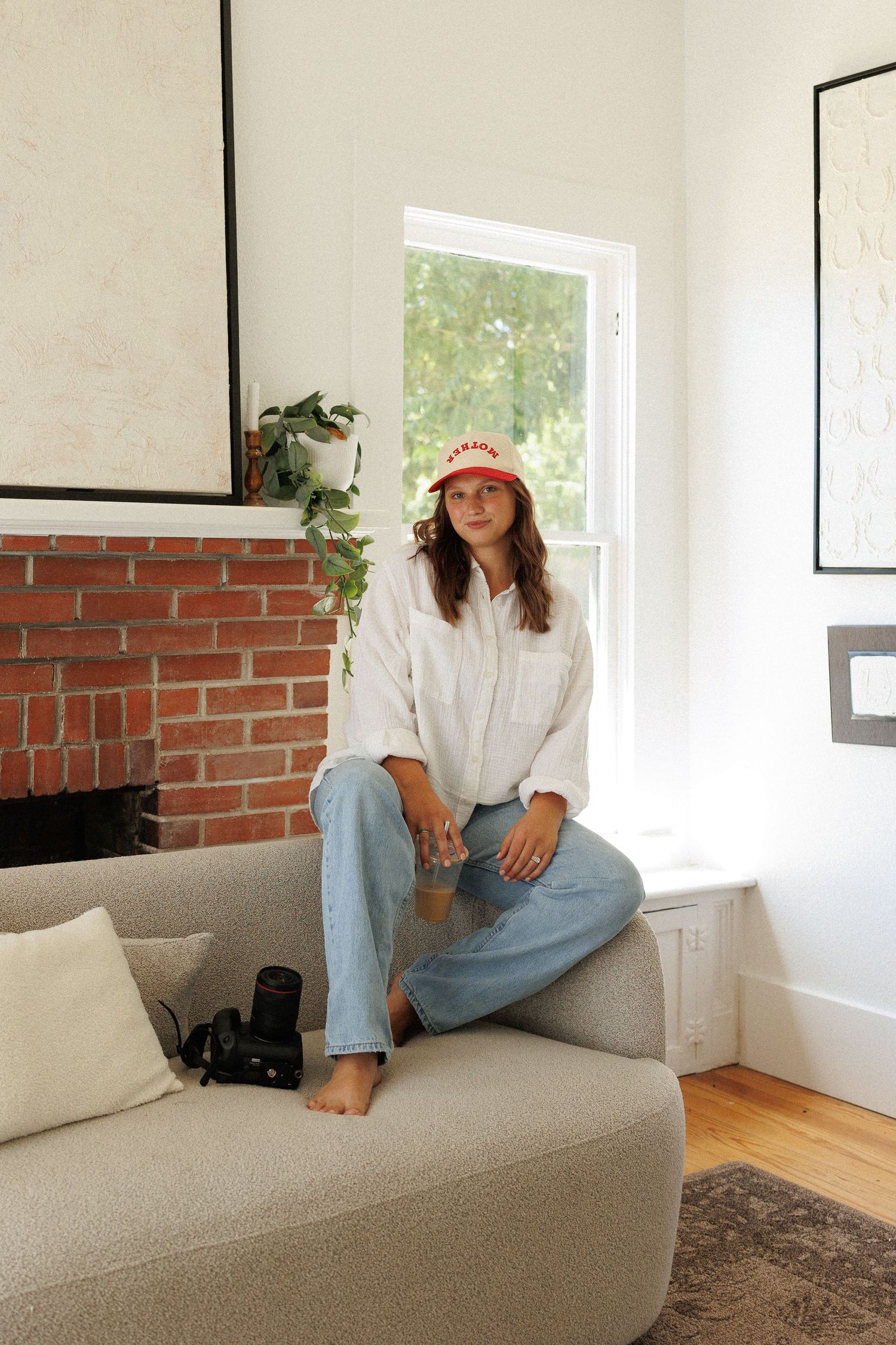 Woman in white shirt, jeans, and red bucket hat sits on gray sofa near brick fireplace in bright living room.