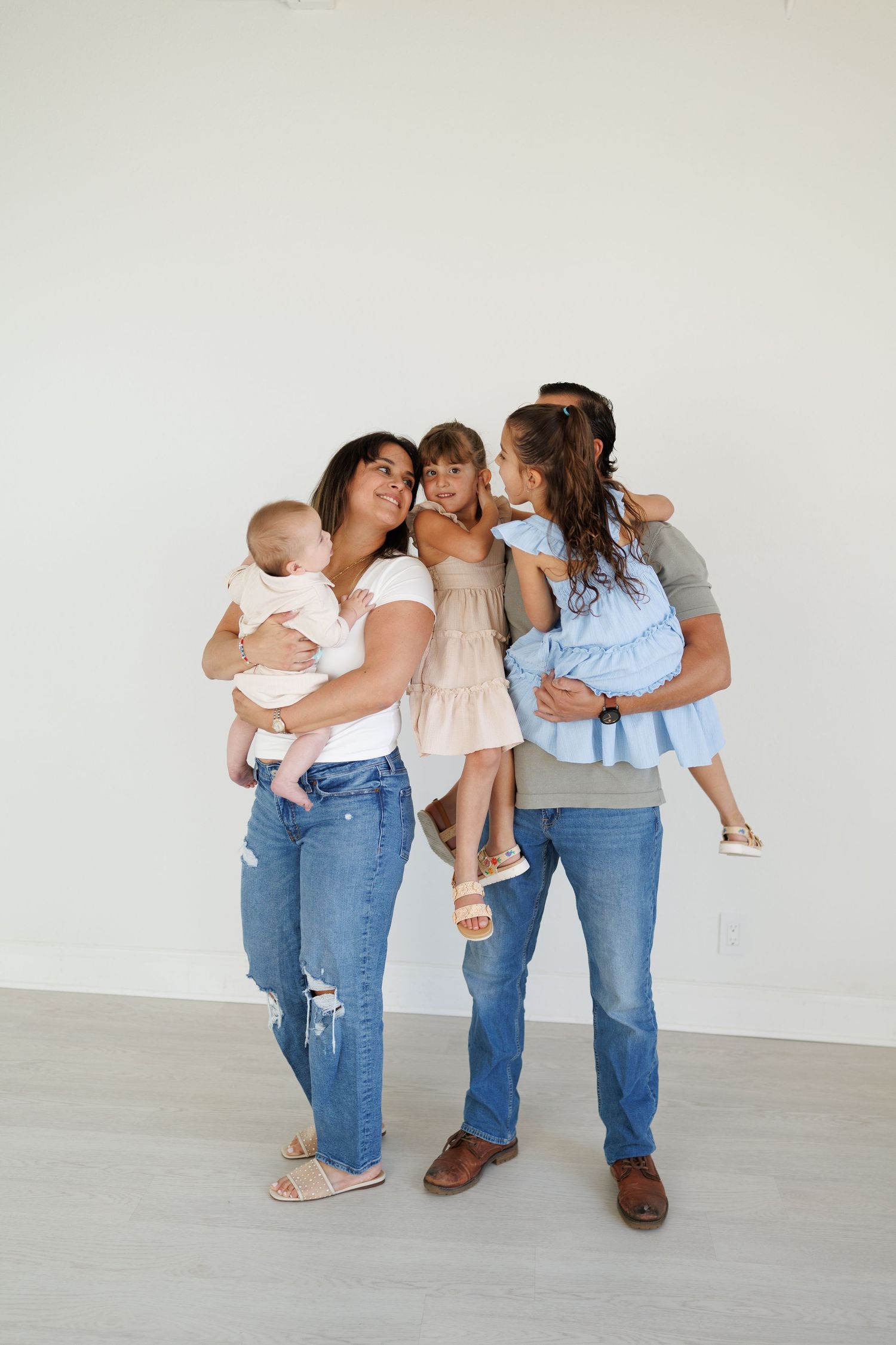 Family wearing casual outfits of jeans and t-shirts stand together against white wall while holding young children.