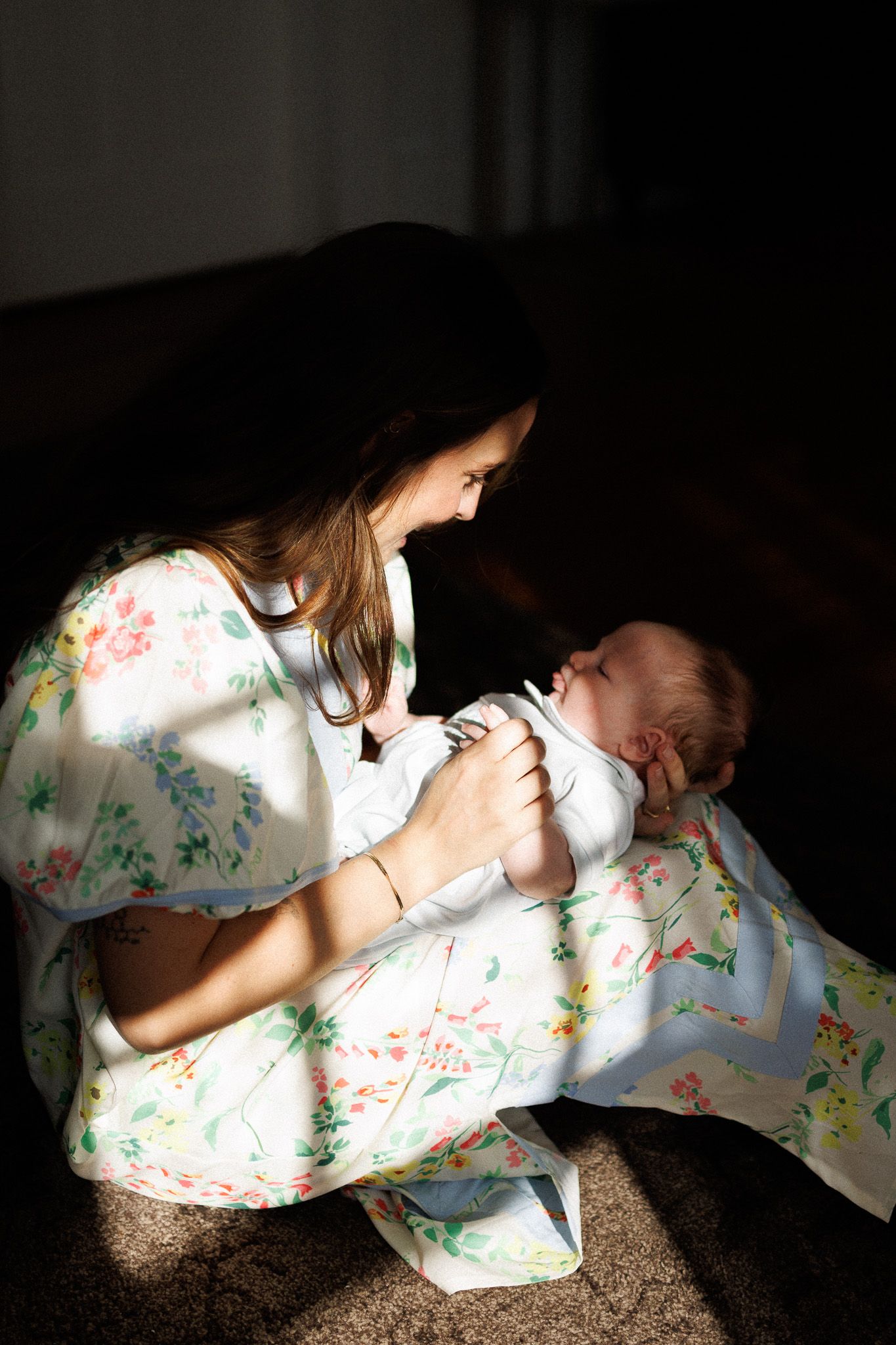 Mother in floral dress tenderly holding and smiling at her newborn baby in warm sunlight.