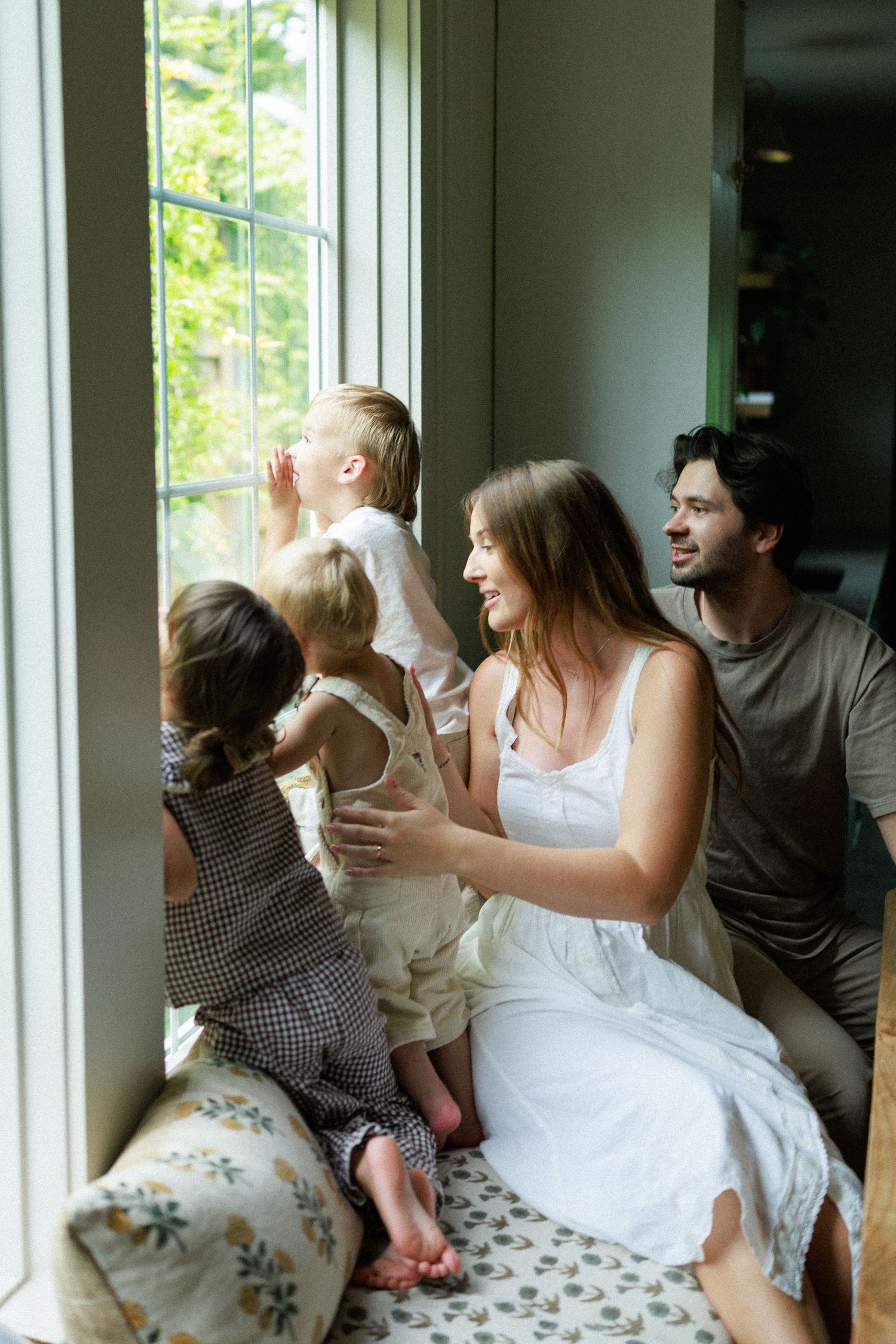 A happy family with two young children sits by a large window, gazing outside in a cozy, light-filled home.